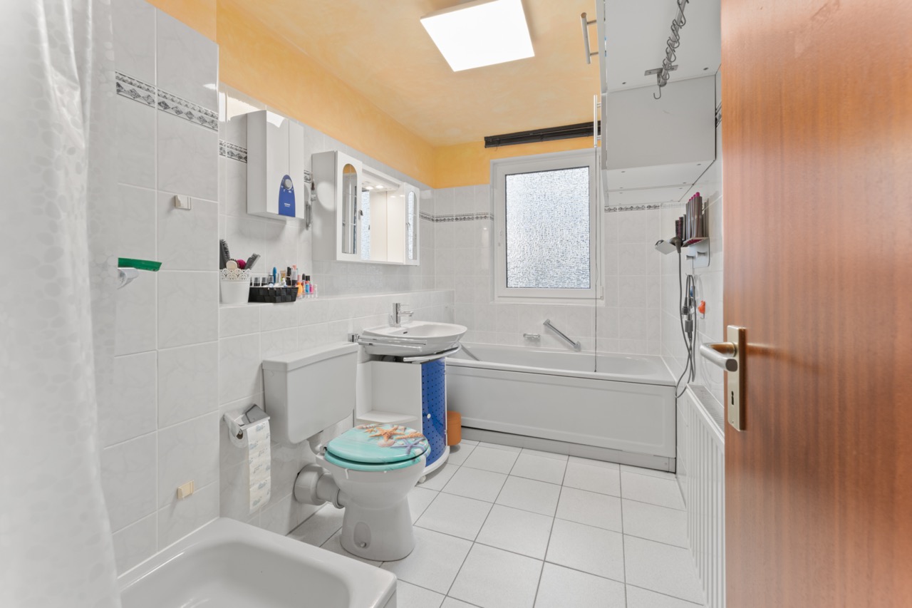 Helles Badezimmer mit Badewanne, Toilette, Waschbecken, Spiegel und Fenster.