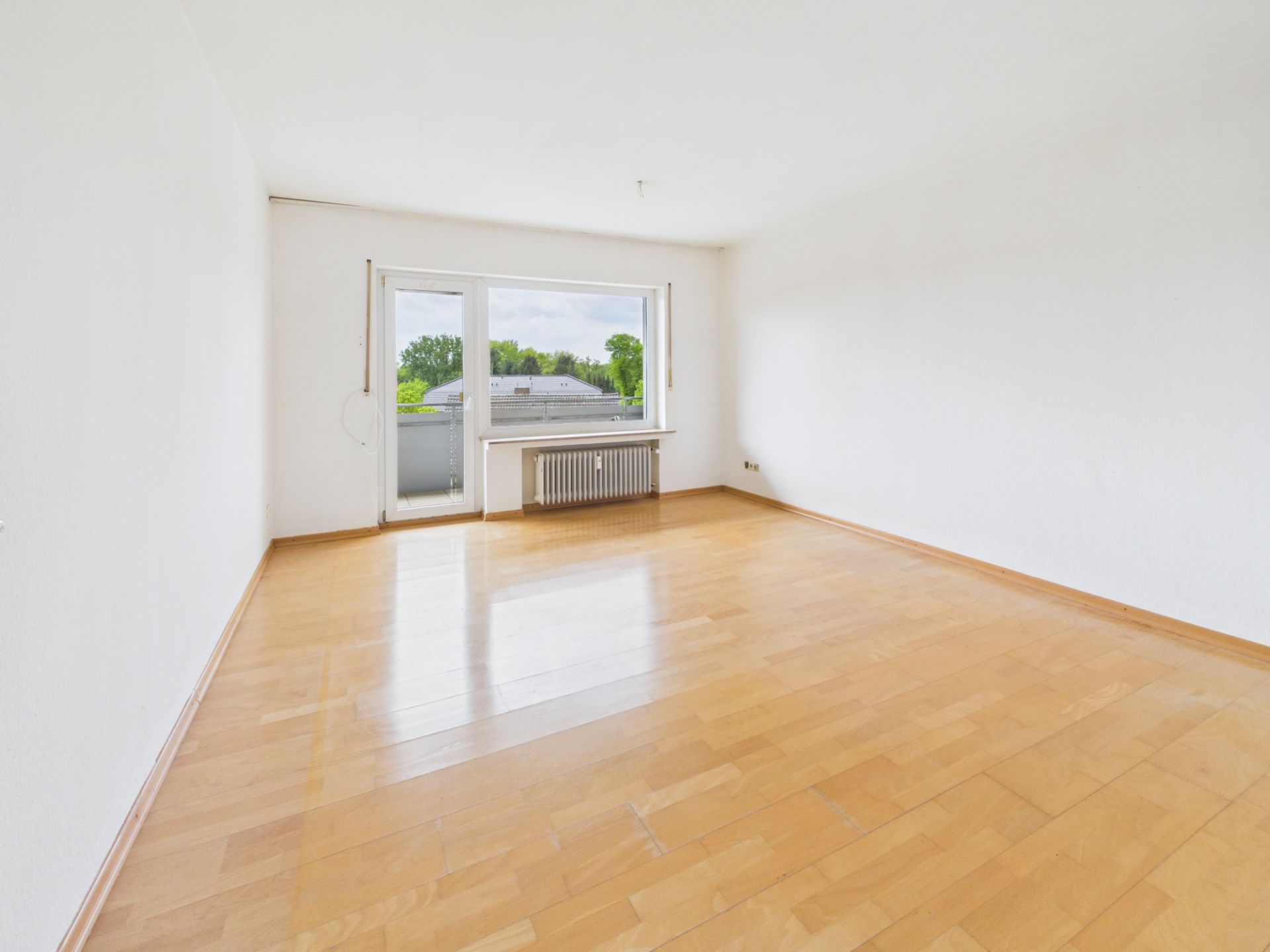 Helles, leeres Zimmer mit Parkettboden, großem Fenster und Zugang zum Balkon.