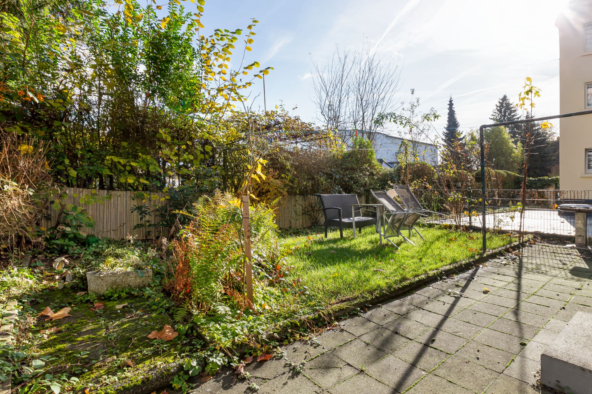 Kleiner Garten mit Terrasse, Gartenmöbeln und viel Grün, umgeben von einem Zaun und Pflanzen.