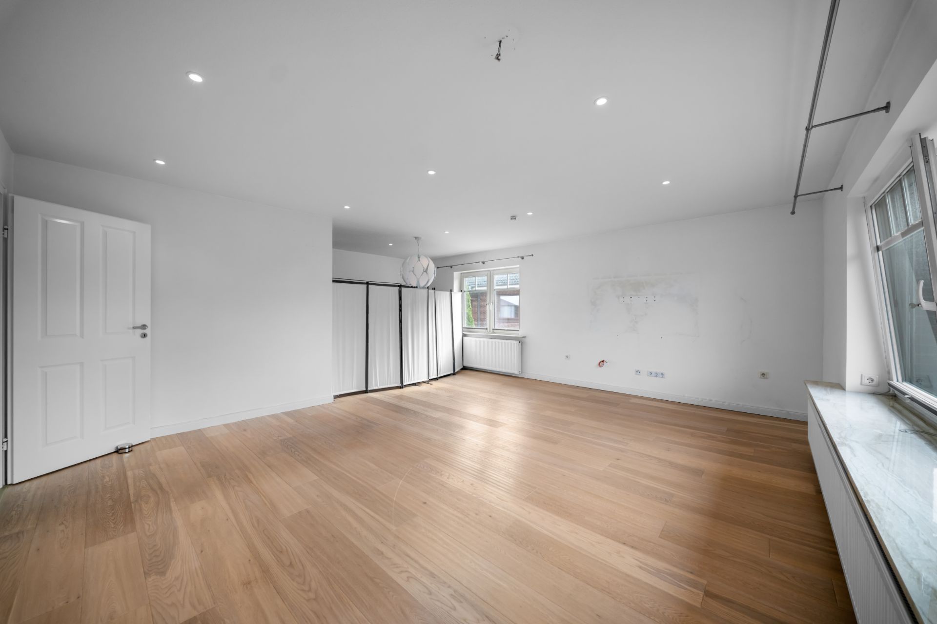 Helles, leeres Zimmer mit Holzboden, weißen Wänden und mehreren Fenstern.
