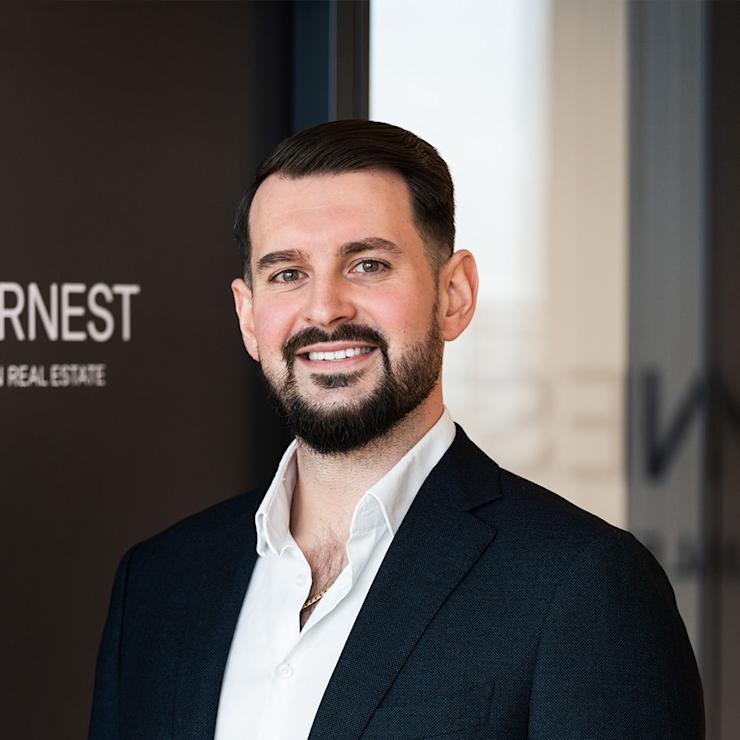 Ein Mann im dunklen Anzug steht vor einer Glastür und lächelt in die Kamera.