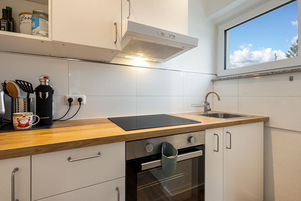 Moderne Küche mit Herd, Spüle, weißen Schränken und Holzarbeitsplatte, daneben ein Fenster.