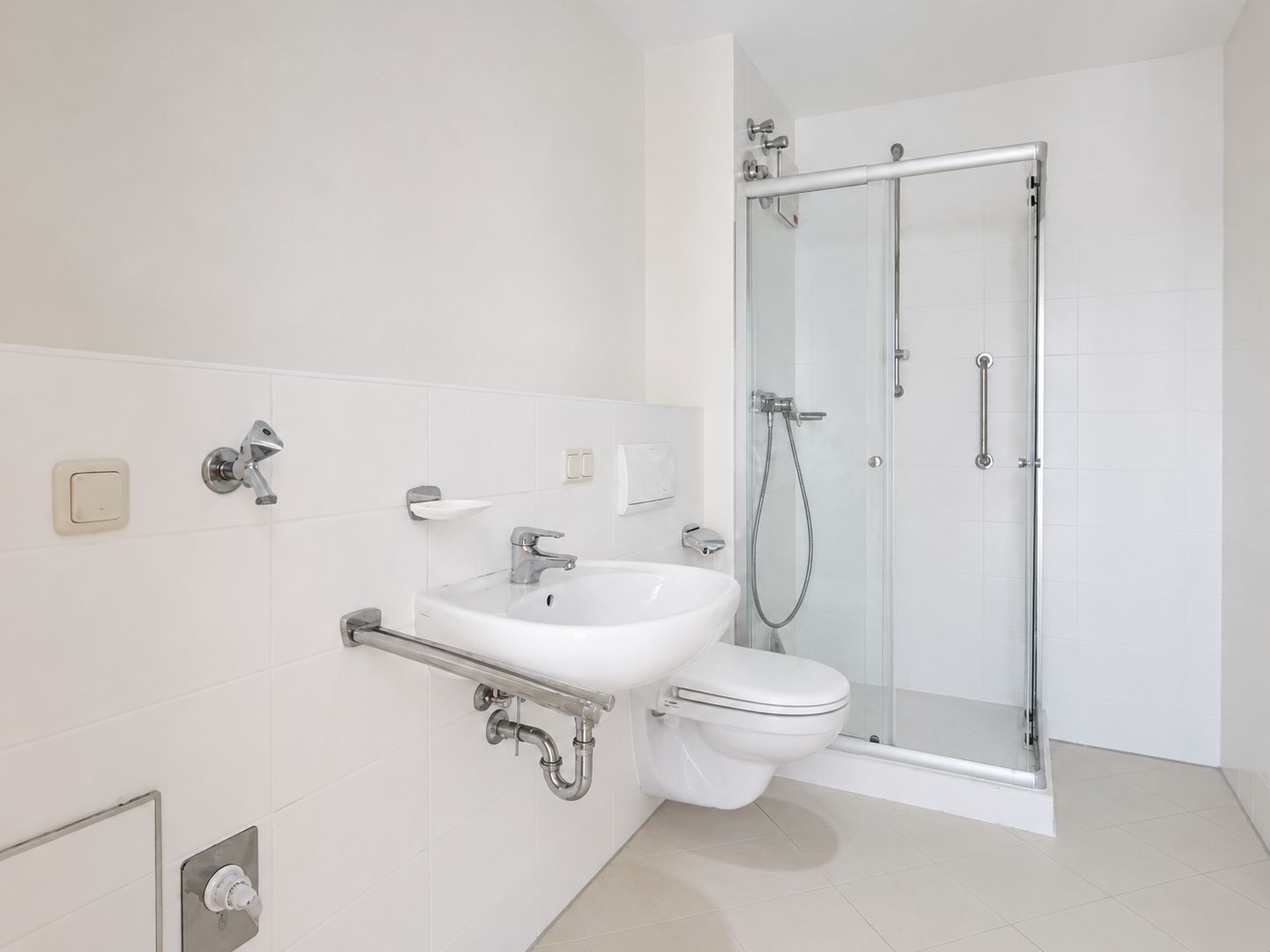 Helles Bad mit Dusche, Wandwaschbecken, Wandtoilette und Haltegriffen.