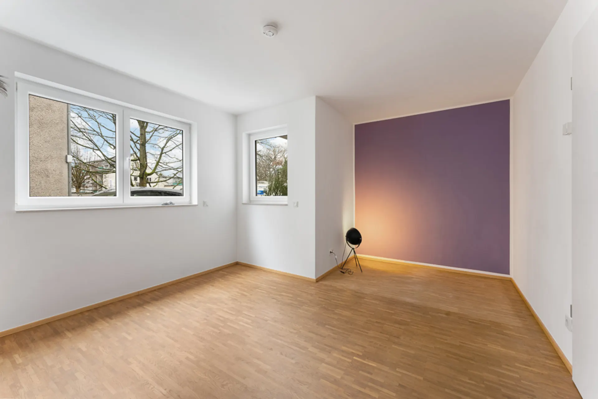 Leerer Raum mit Holzfußboden, großen Fenstern und lila Wand, Stehlampe in der Ecke.