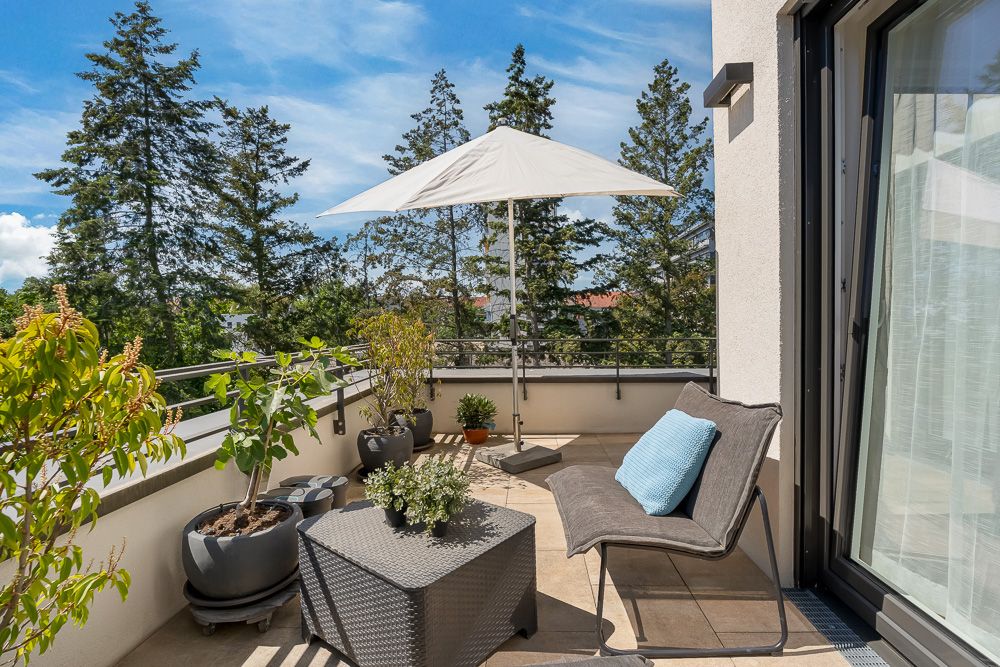 Balkon mit Gartenmöbeln, Sonnenschirm, Pflanzen und Blick auf Bäume bei sonnigem Wetter.