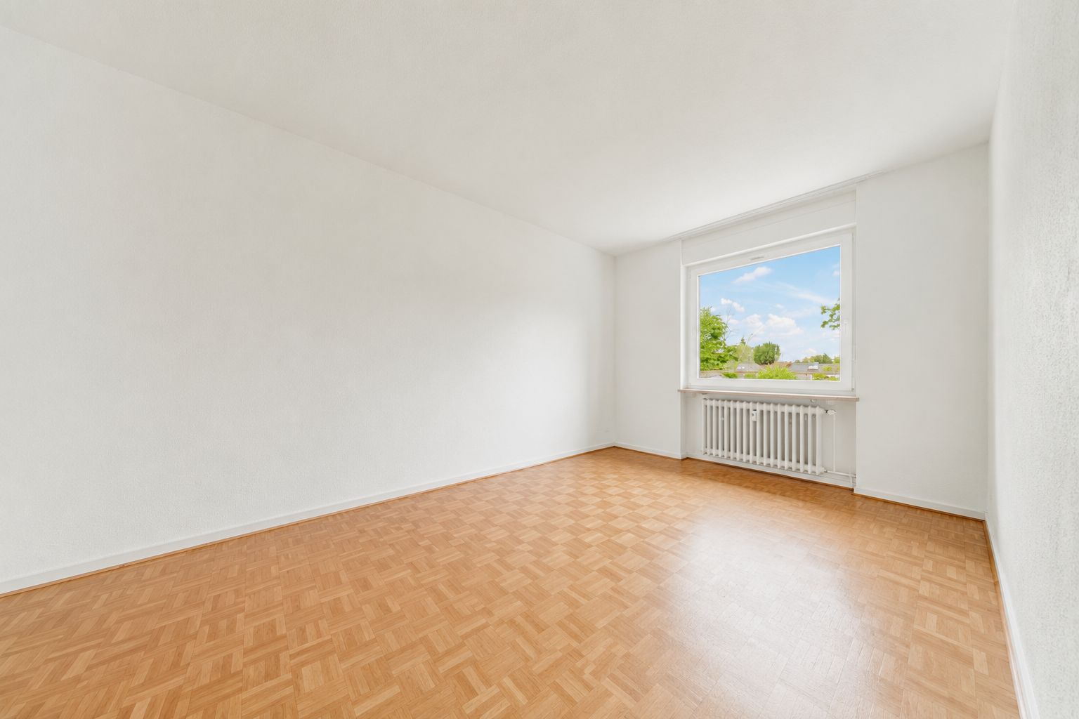 Leerer, heller Raum mit Parkettboden und großem Fenster mit Blick nach draußen.
