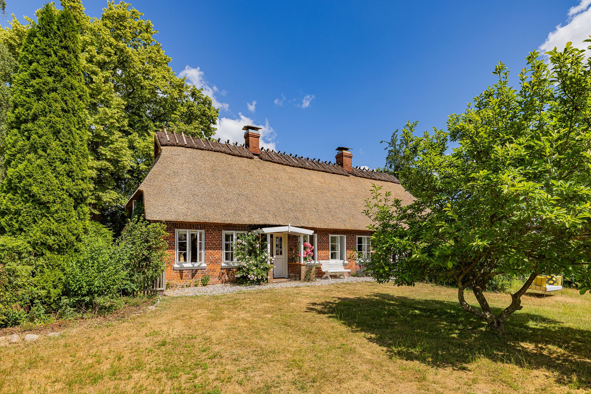 Backsteinhaus mit Reetdach, umgeben von Garten, Bäumen und blühenden Sträuchern bei sonnigem Wetter.