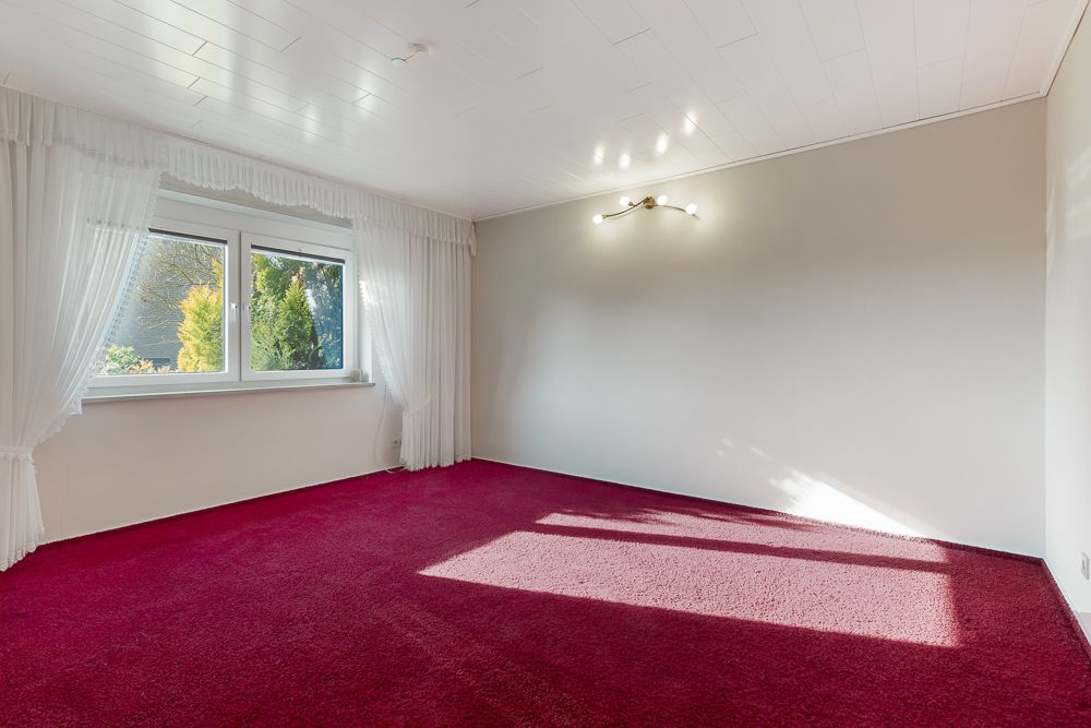Leeres Zimmer mit roten Teppichboden, weißen Wänden, großen Fenstern und Deckenleuchte.