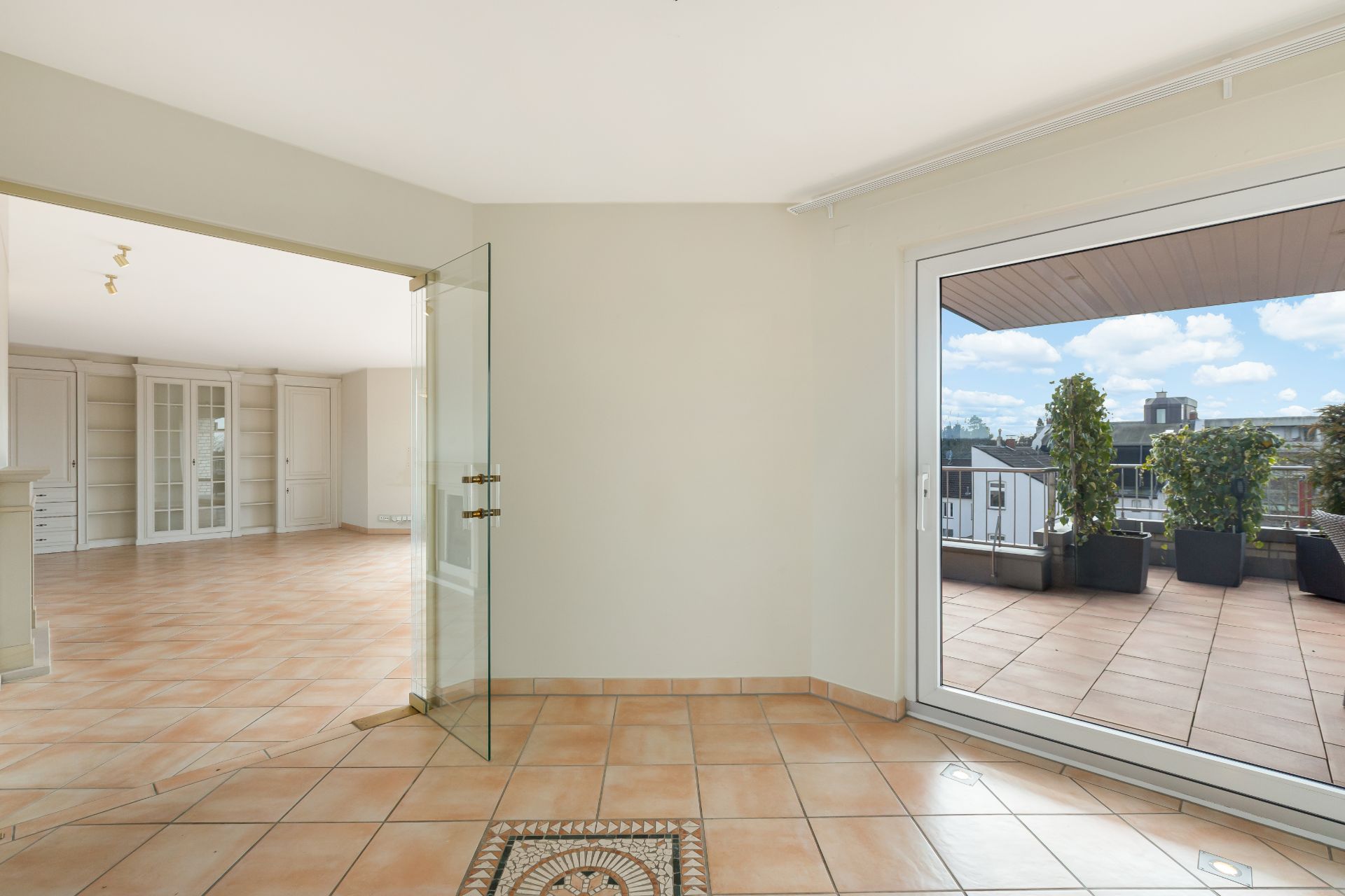Helles Wohnzimmer mit Fliesenboden, Glastür und Zugang zu einer Terrasse mit Pflanzen.