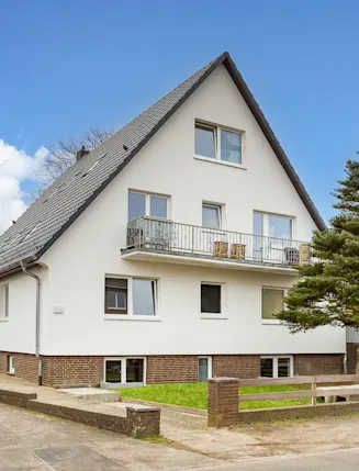 Zweistöckiges Wohnhaus mit Satteldach, Balkon und Garten, große Tanne daneben.