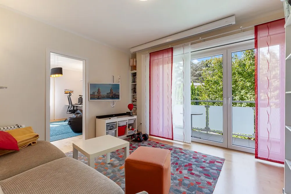 Helles Wohnzimmer mit Sofa, Tisch, Teppich und Zugang zum Balkon mit Blick auf Bäume.