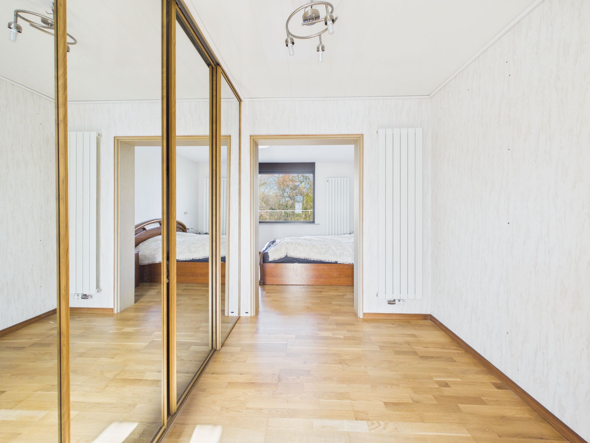 Zimmer mit Holzfußboden, Spiegel-Schiebetüren und Blick auf ein Schlafzimmer mit Fenster.