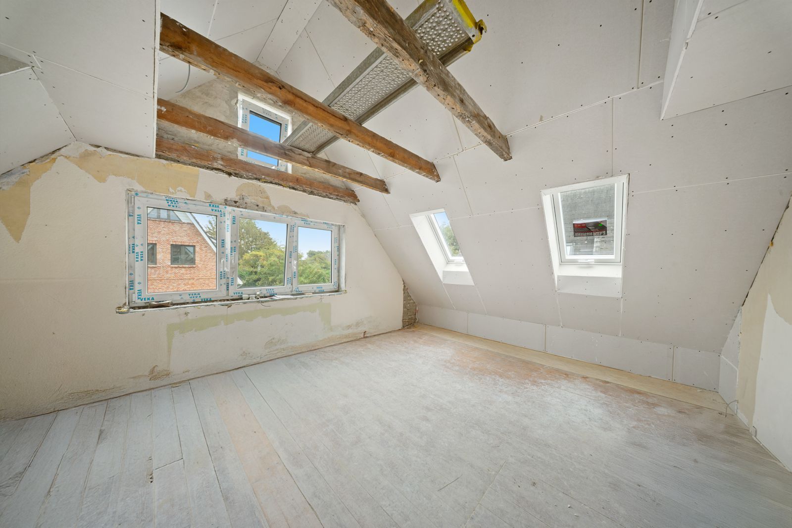Helles Dachgeschosszimmer mit sichtbaren Holzbalken und mehreren Fenstern im Ausbauzustand.