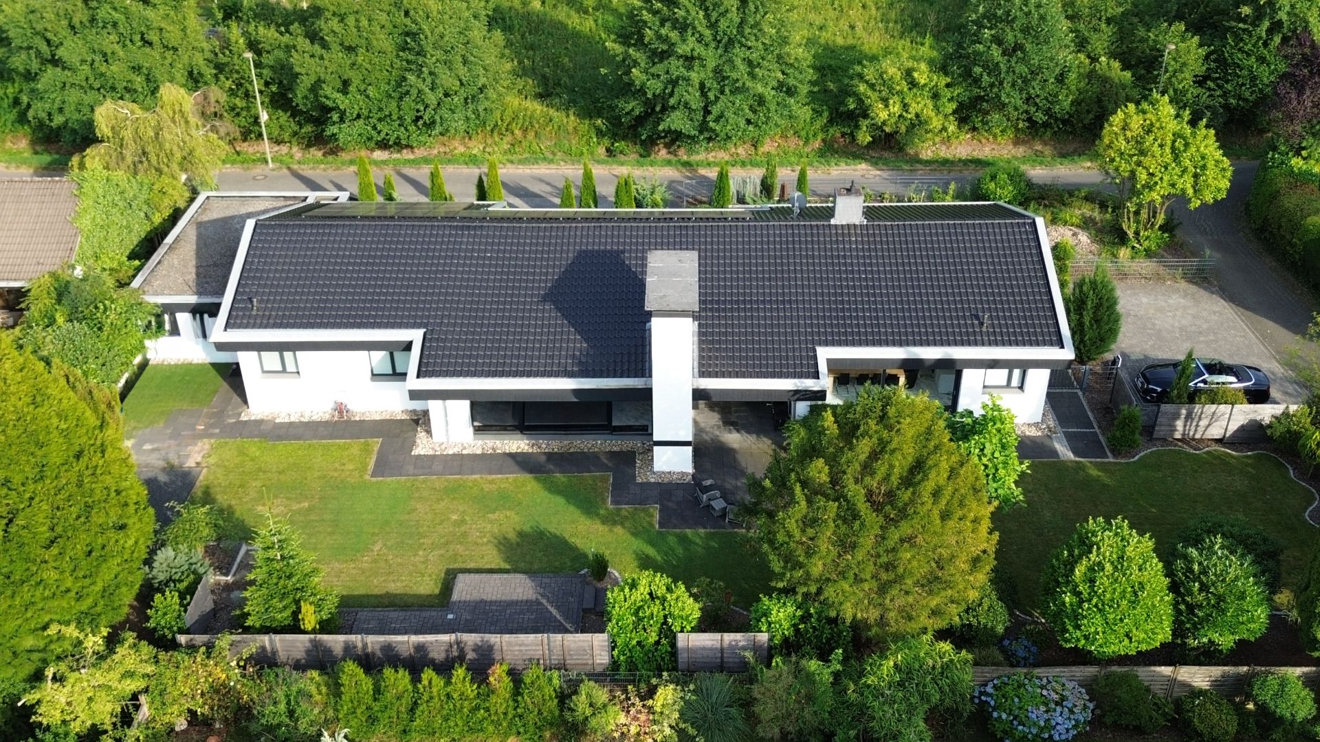 Moderne, einstöckige Wohnhaus mit schwarzem Dach, umgeben von Garten und Bäumen aus der Vogelperspektive.