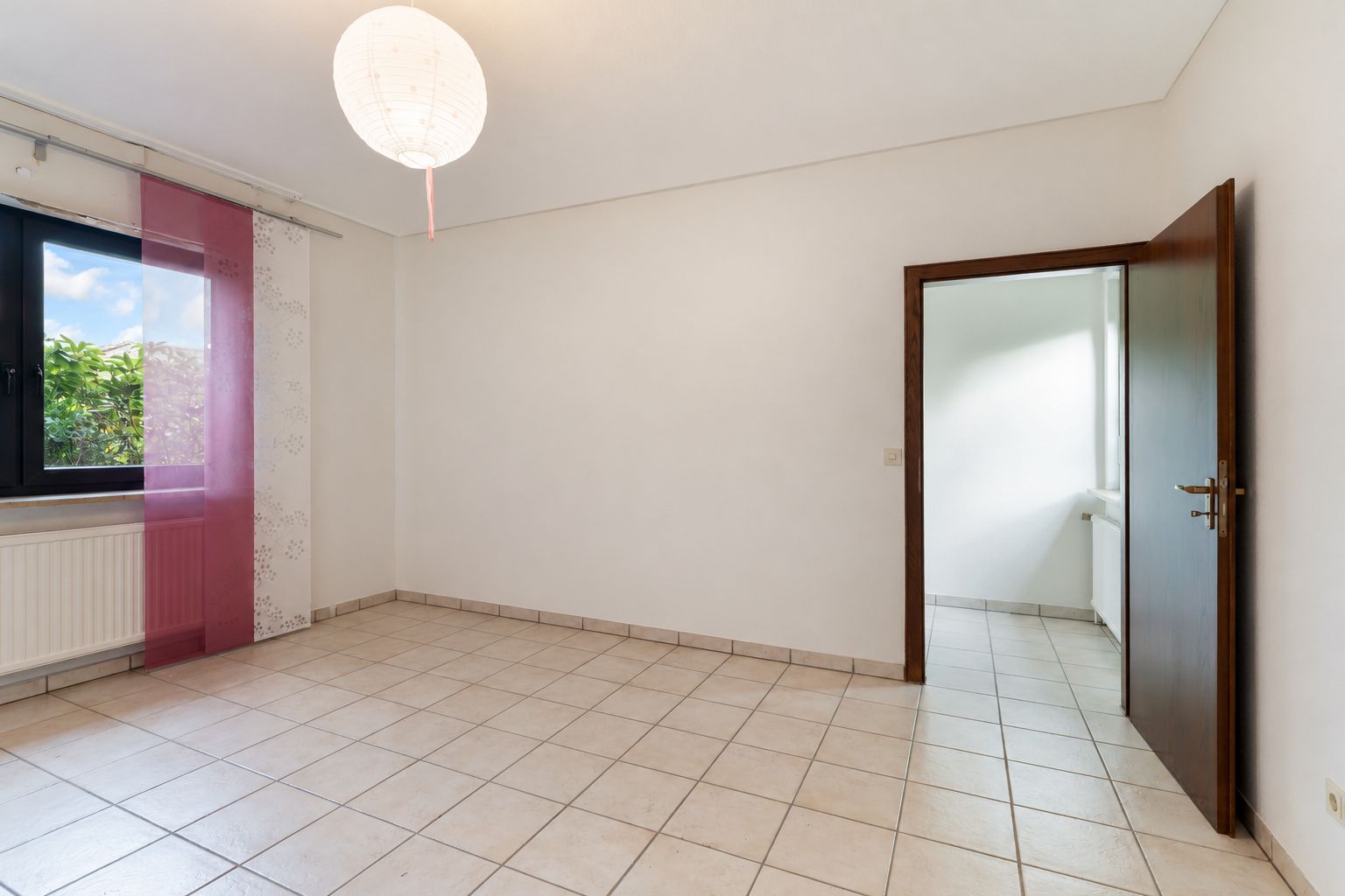 Leeres Zimmer mit Fliesenboden, offenem Fenster, rosa Vorhang und offener Tür.