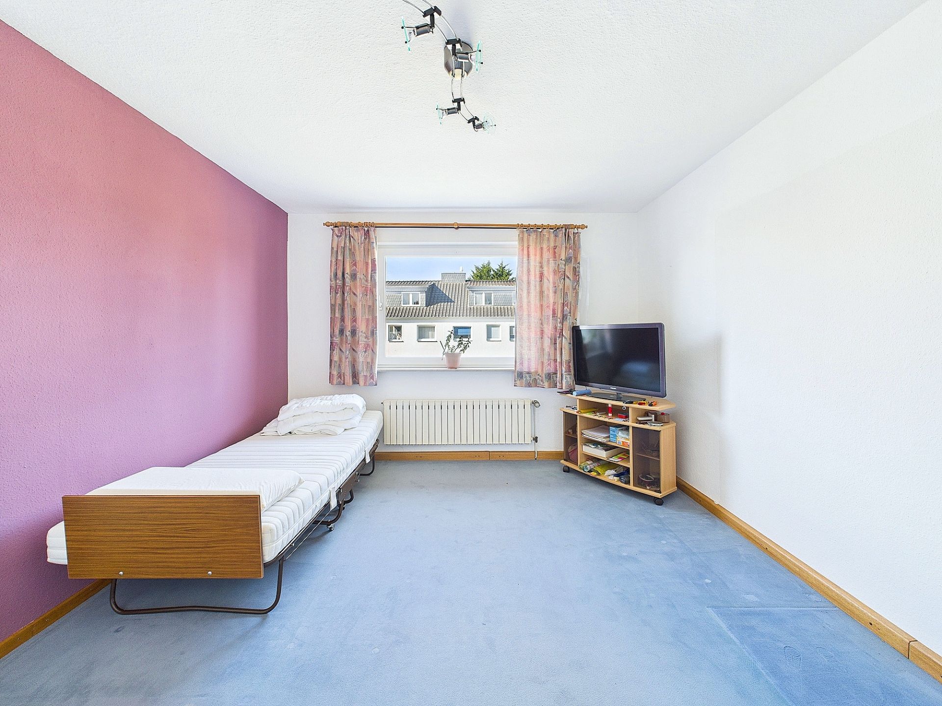 Helles Zimmer mit Einzelbett, Fernseher und großem Fenster mit Vorhängen.