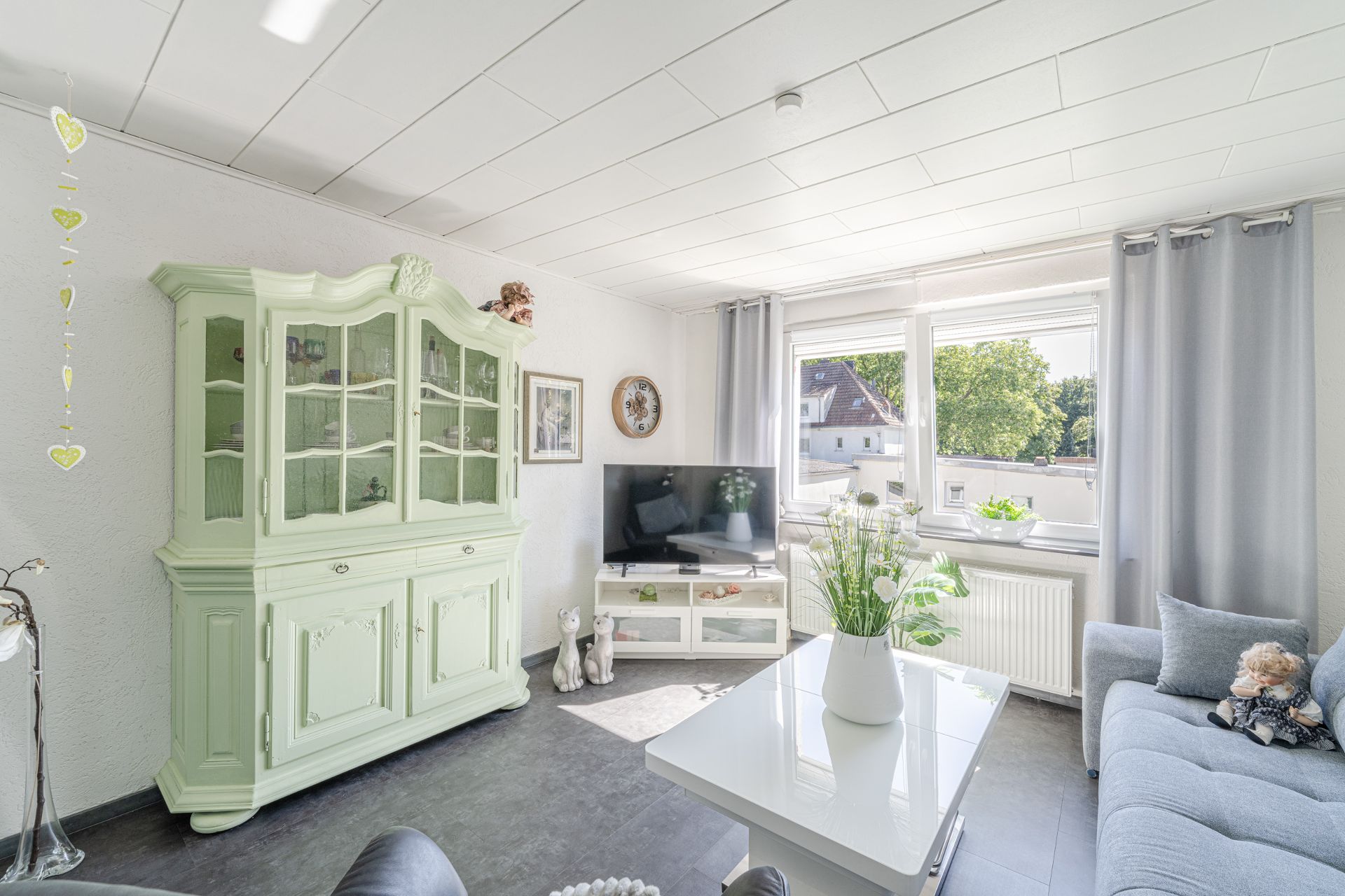 Helles Wohnzimmer mit grüner Vitrine, grauem Sofa, Fernseher und großem Fenster.