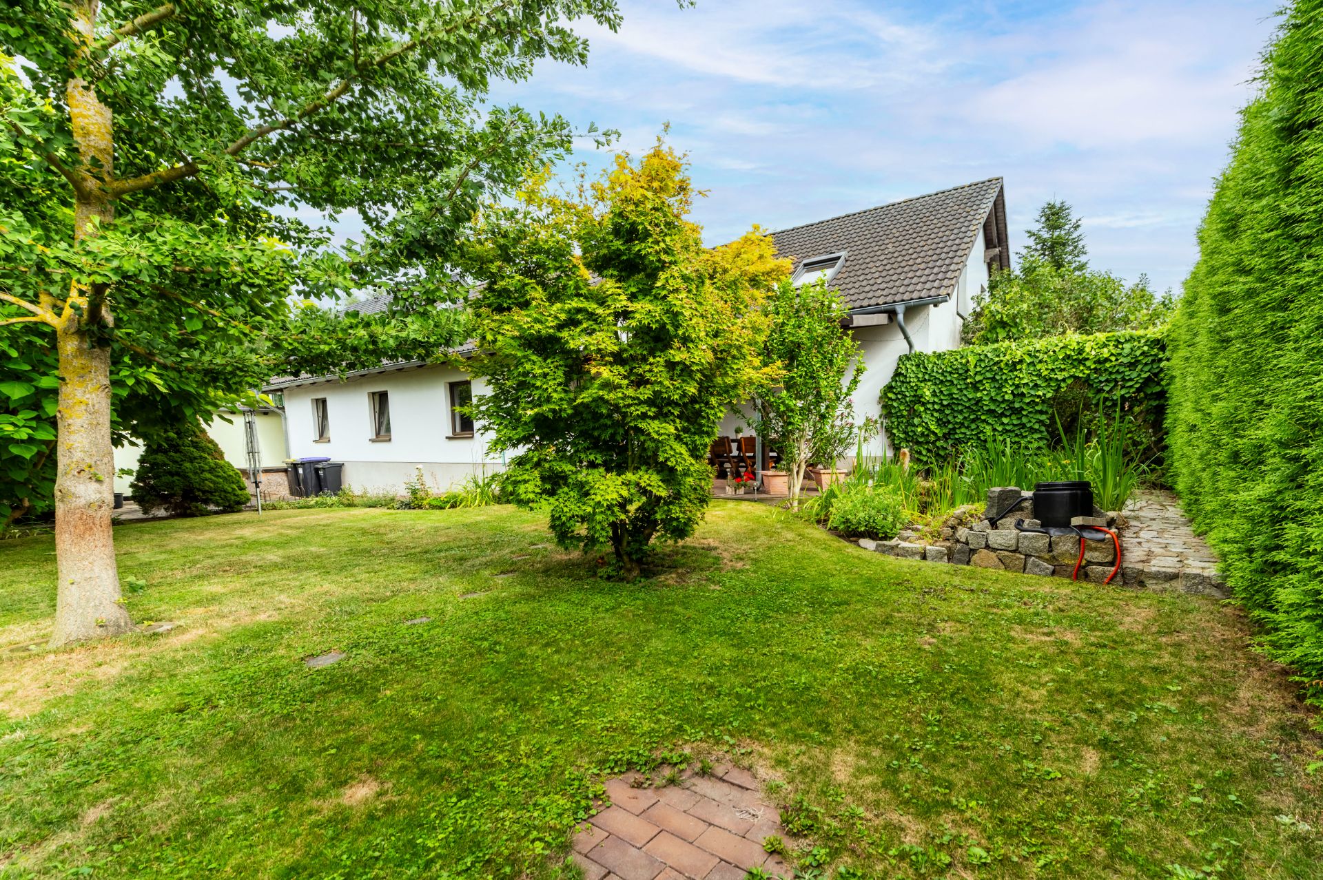 Garten mit Rasen, Bäumen, Sträuchern und Blick auf ein weißes Haus mit Terrasse.