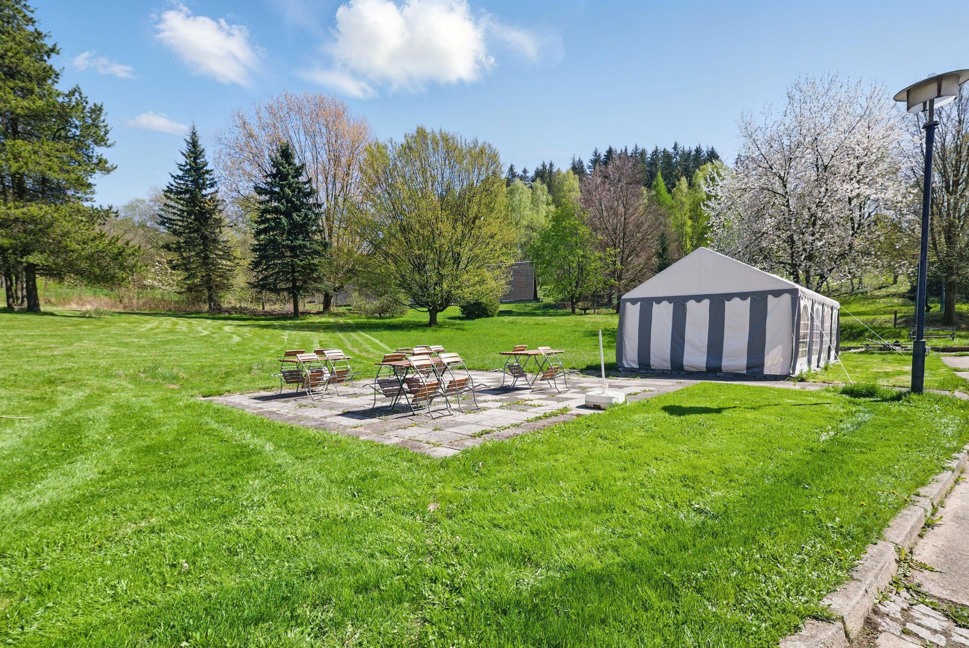 Garten mit Zelt, mehreren Tischen und Stühlen auf einer Steinfläche, umgeben von Bäumen.