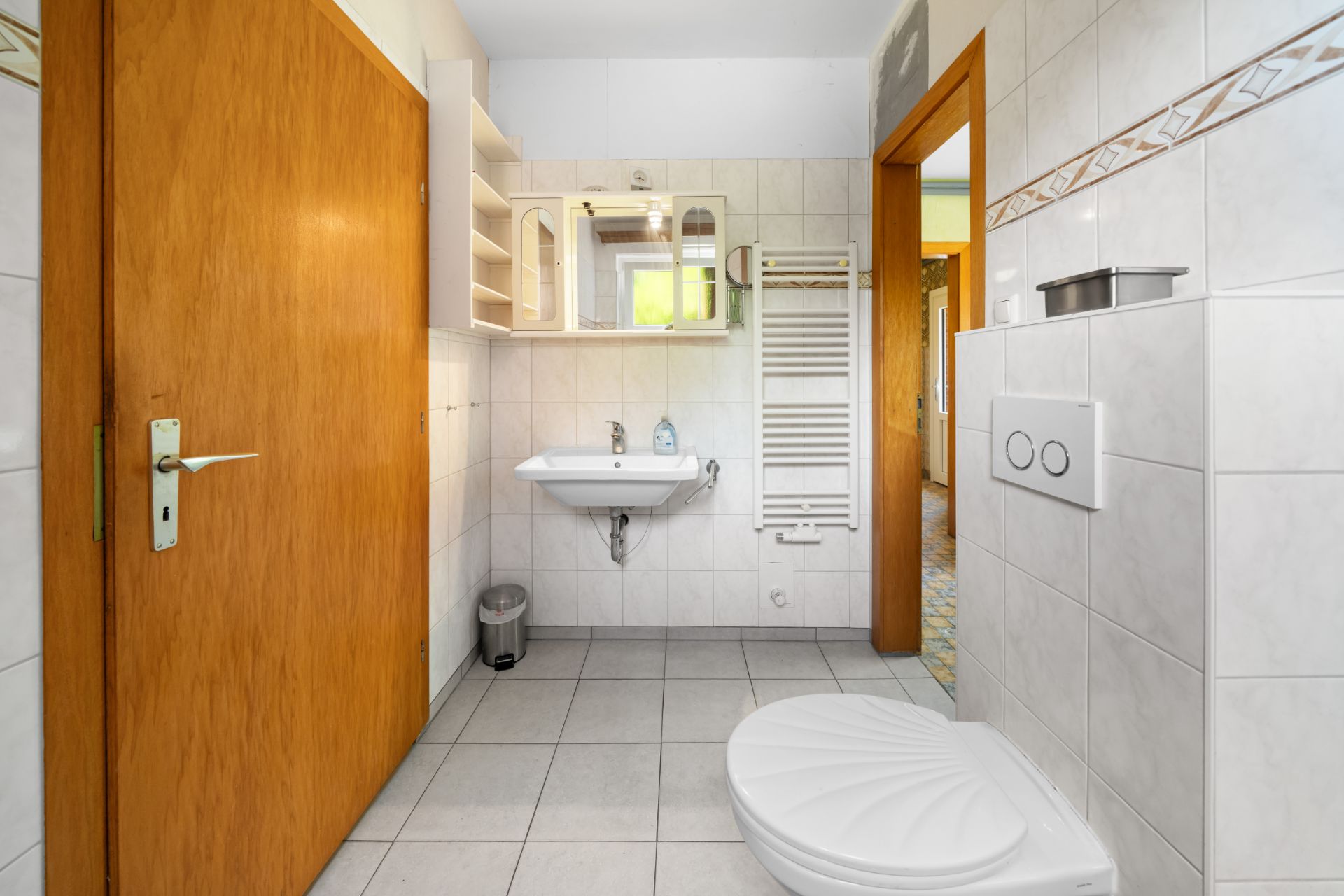 Helles Badezimmer mit Wandwaschbecken, Spiegel, Heizkörper und Toilette.