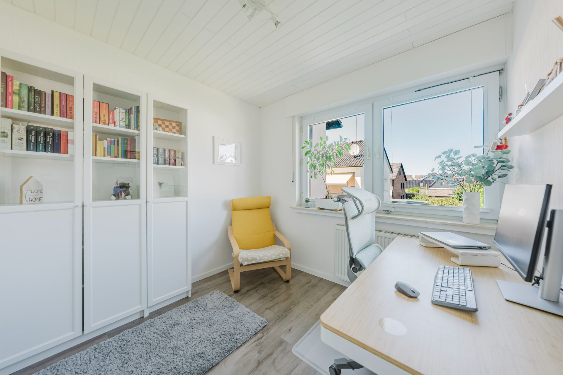 Helles Arbeitszimmer mit Schreibtisch, gelbem Sessel, Bücherregalen und großem Fenster.