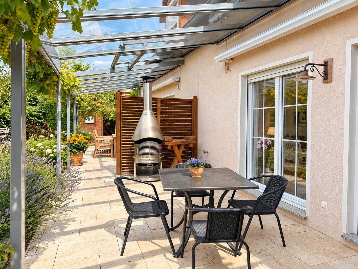 Überdachte Terrasse mit Tisch, vier Stühlen, Ofen und Pflanzen neben einem Haus.