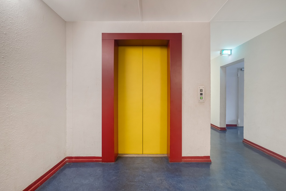 Gelbe Aufzugtür mit rotem Rahmen in einem Flur eines Wohngebäudes.