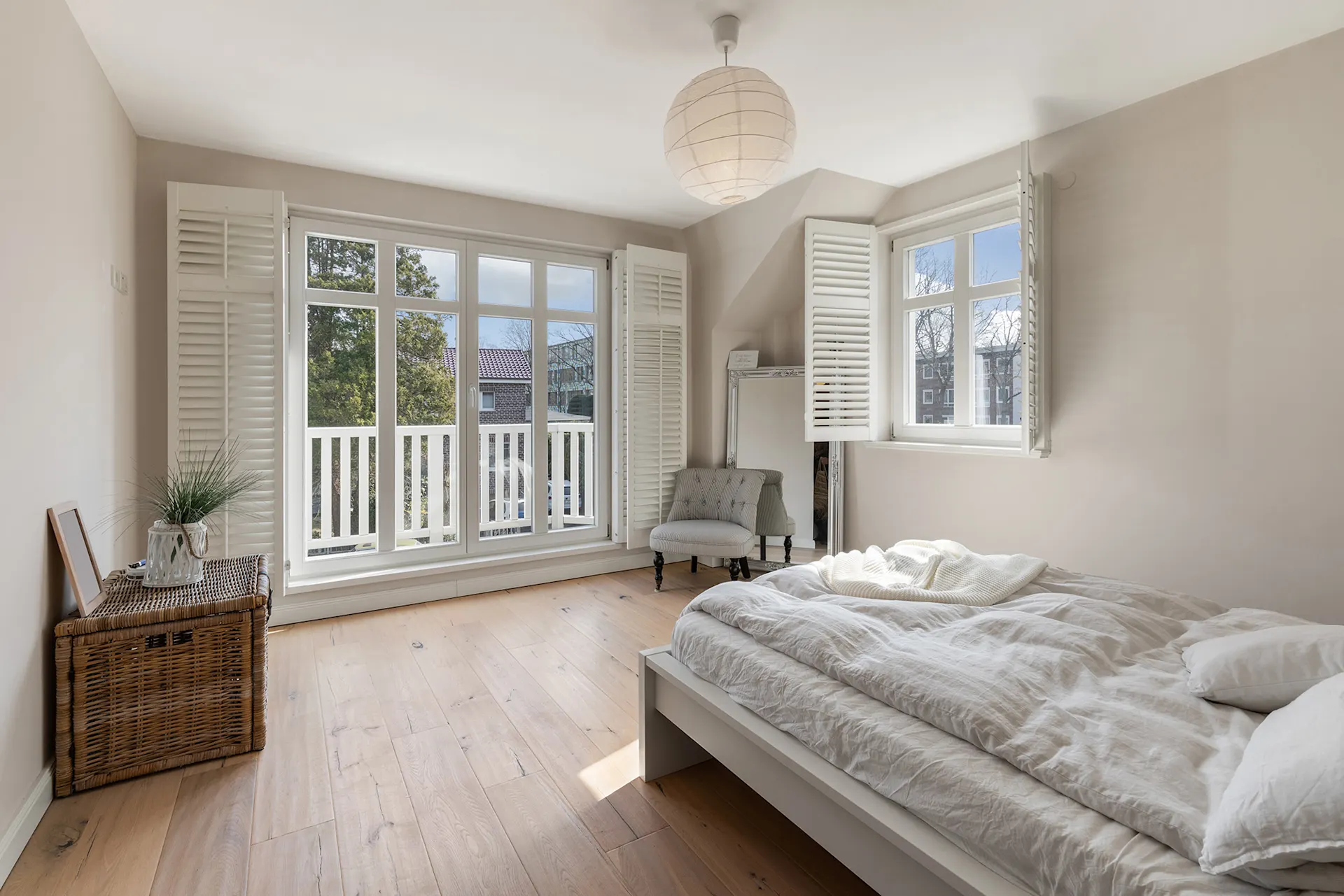 Helles Schlafzimmer mit Doppelbett, Sessel, Korbtruhe und großen Fenstern mit weißen Fensterläden.