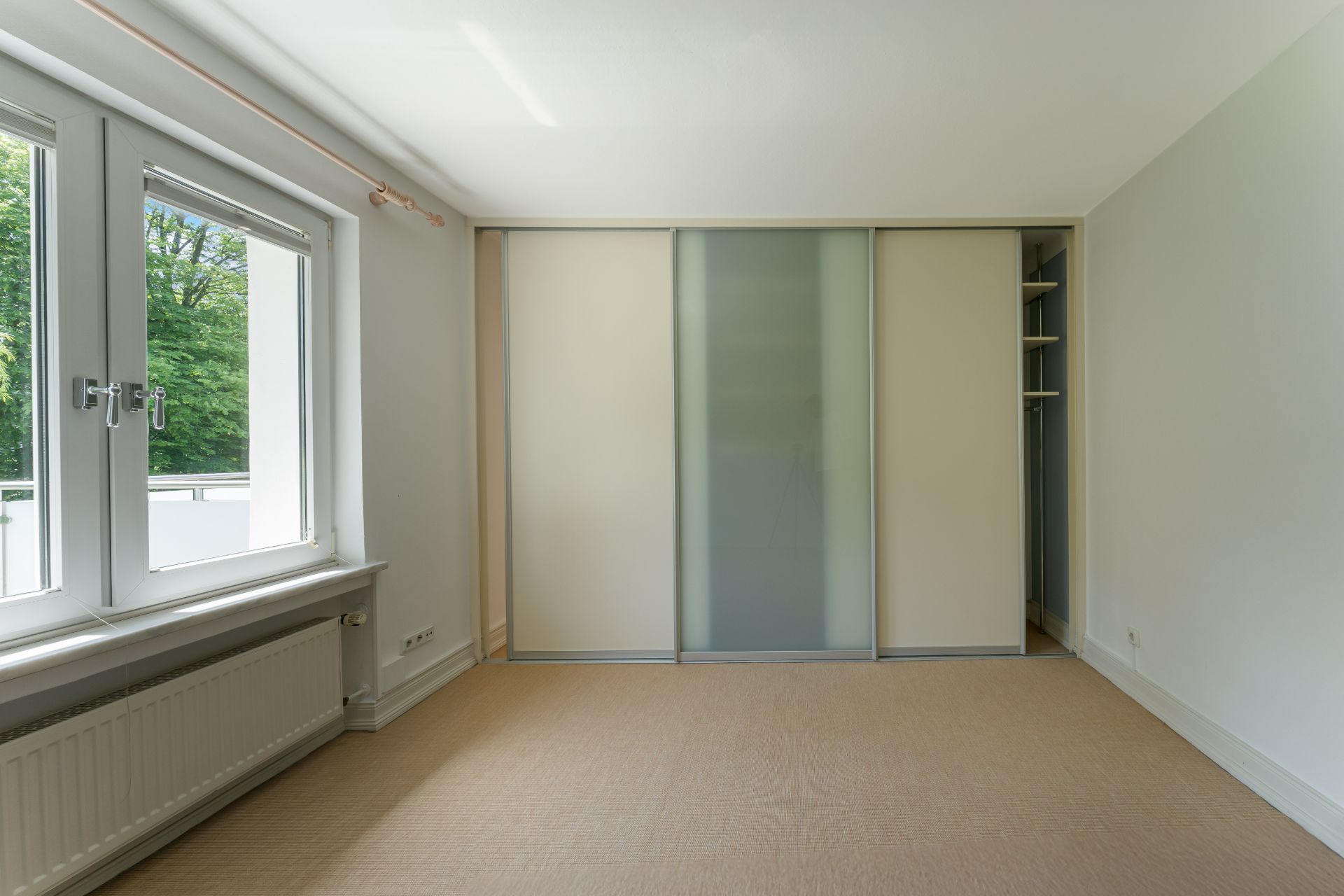 Helles Zimmer mit Teppichboden, großem Fenster und Einbauschrank mit Schiebetüren.