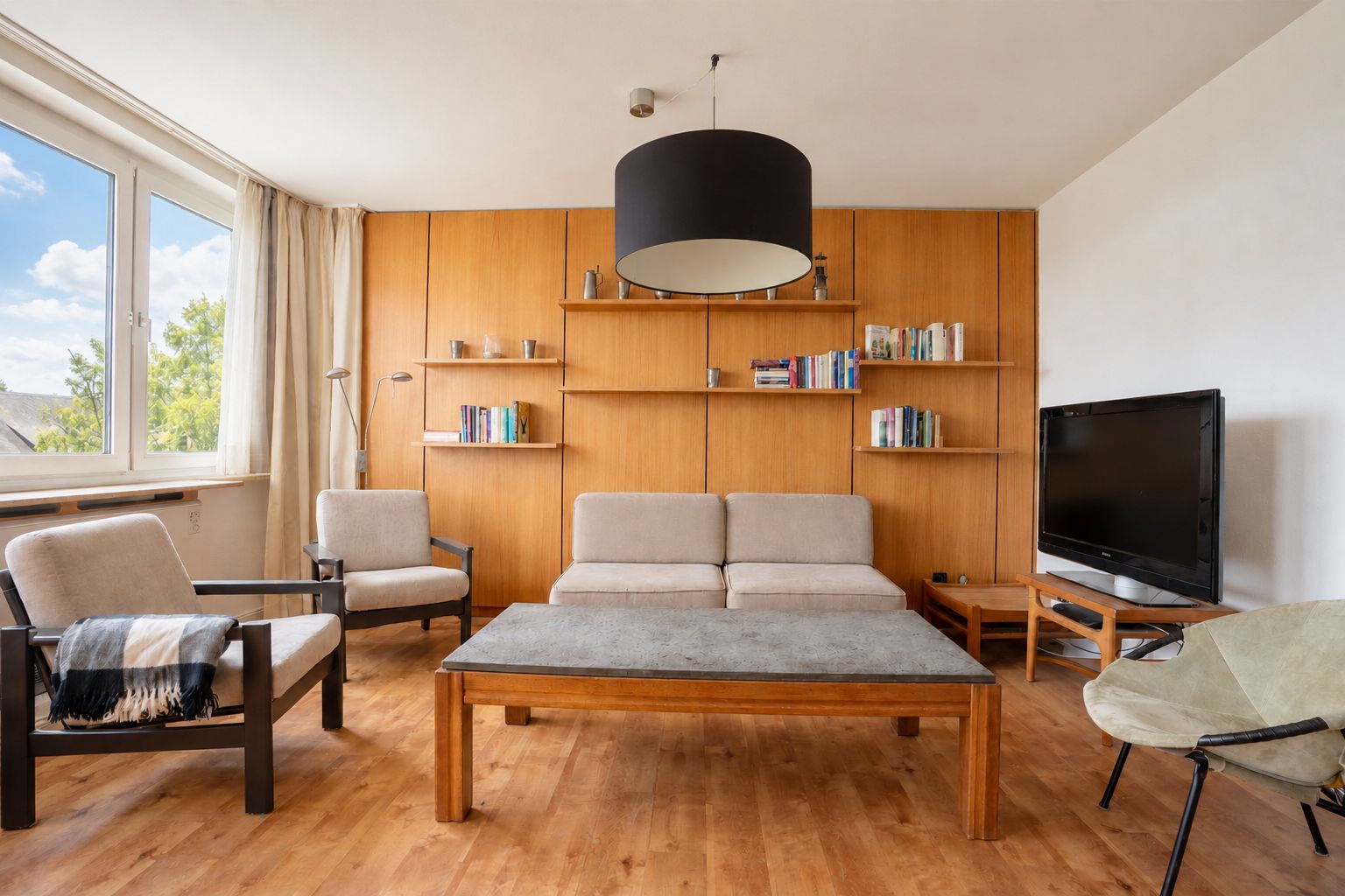 Helles Wohnzimmer mit Sofa, Sessel, Couchtisch, Bücherregal und Fernseher auf Holzfußboden.