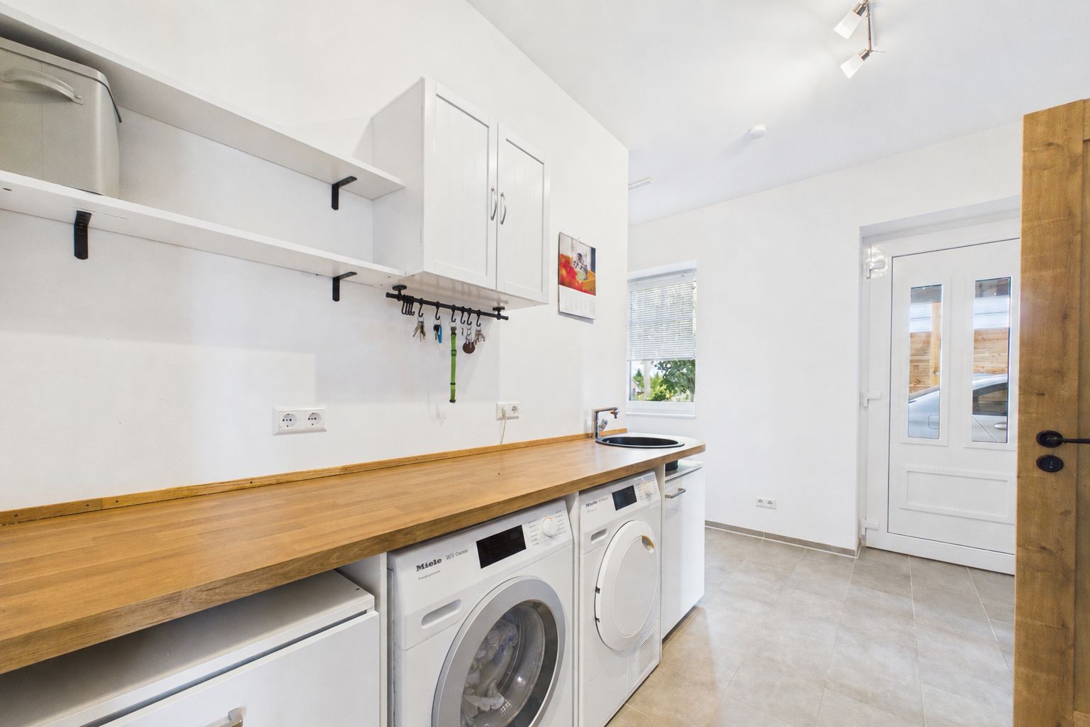 Heller Hauswirtschaftsraum mit Holzarbeitsplatte, Waschmaschine, Trockner und Regalen.