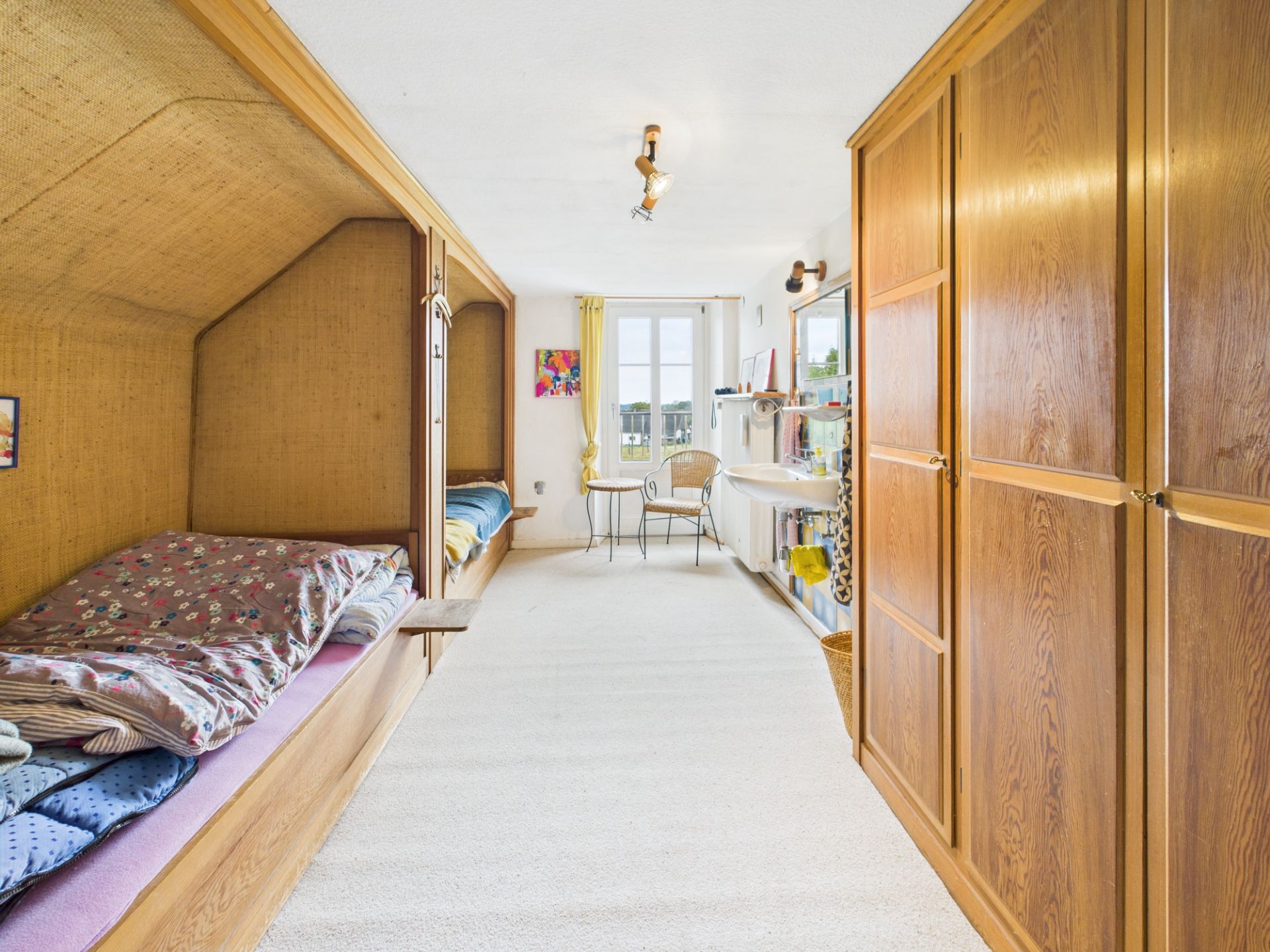 Helles Zimmer mit zwei Holzbettkojen, Kleiderschrank, Waschbecken und Fenster mit Blick nach draußen.