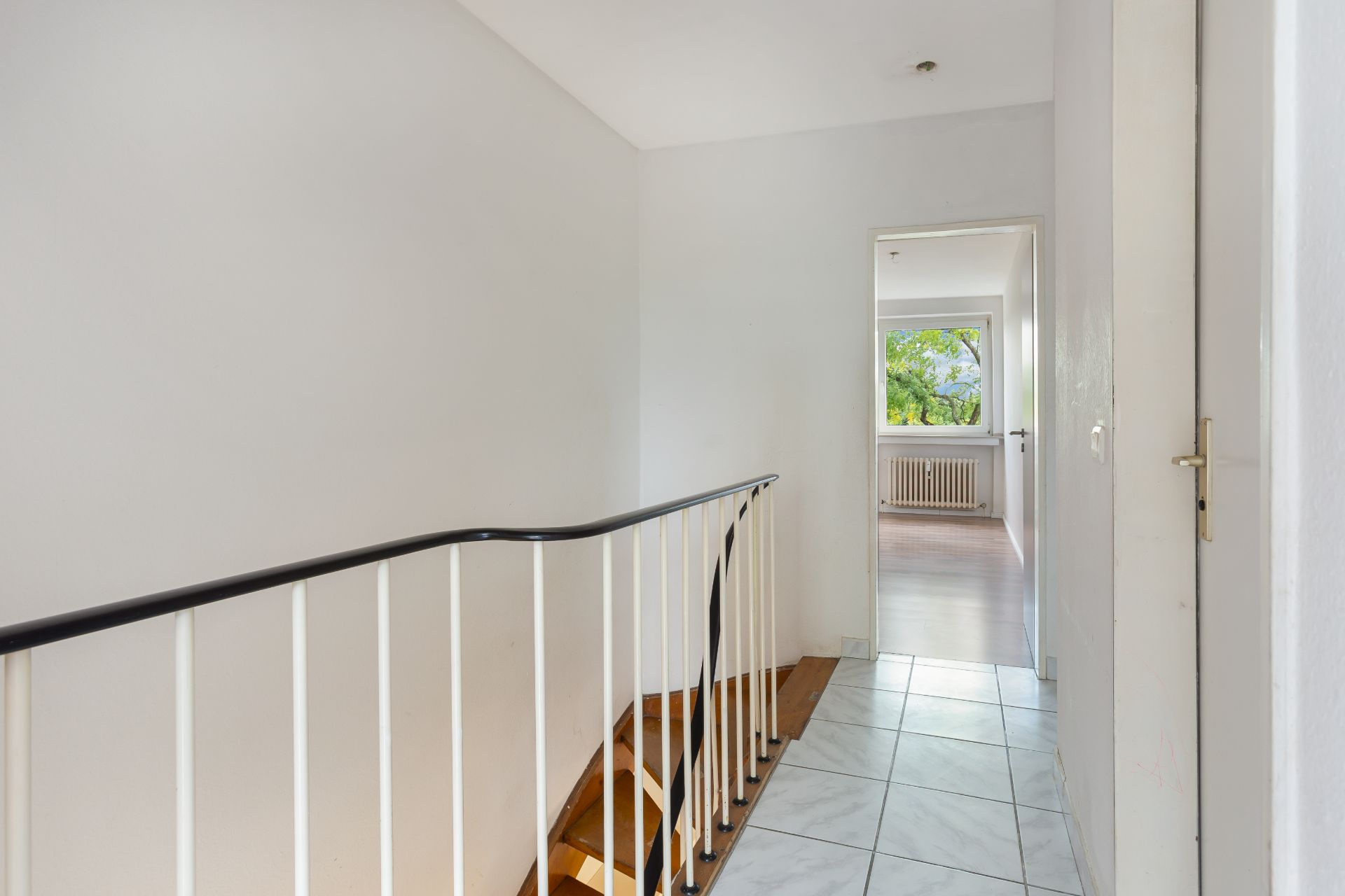 Heller Flur mit Treppe, Fliesenboden und Blick in ein Zimmer mit Fenster und Heizkörper.