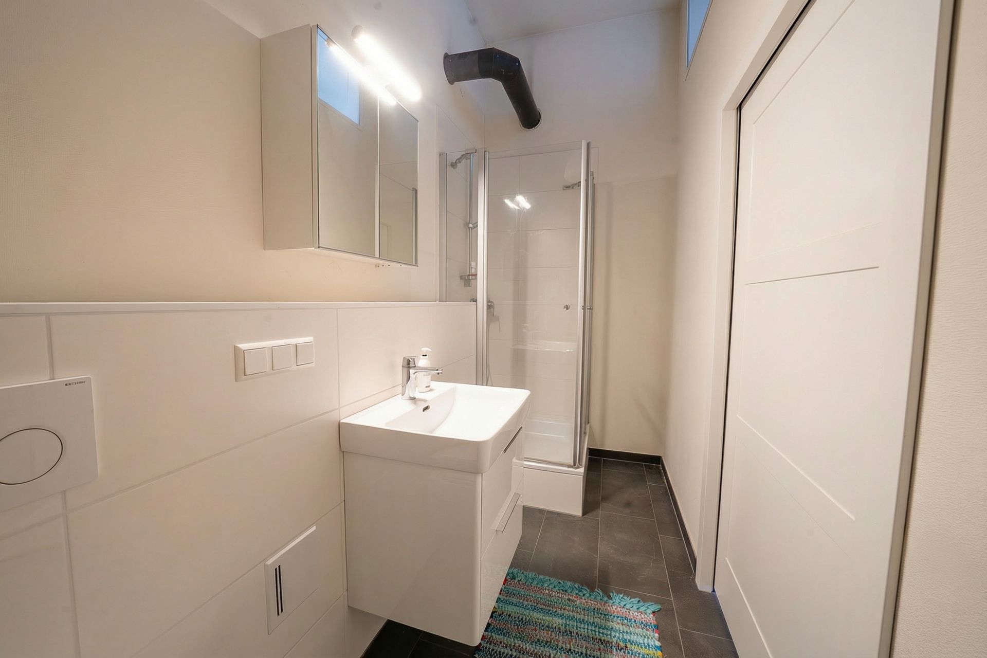 Modernes Badezimmer mit Waschbecken, Spiegel, Dusche und buntem Teppich auf dunklem Boden.