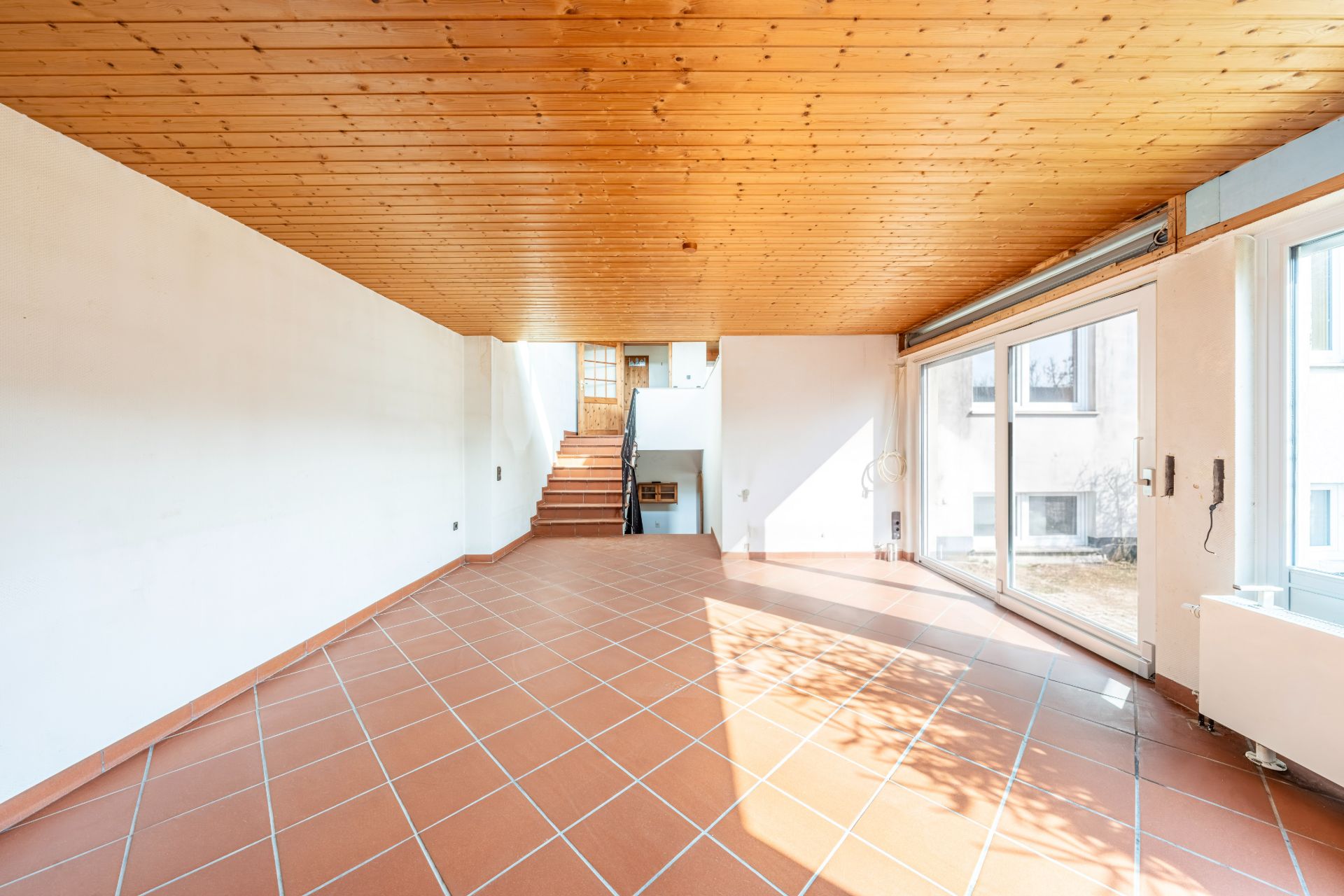 Helles Wohnzimmer mit Fliesenboden, Holzdecke, großen Fenstern und einer Treppe im Hintergrund.