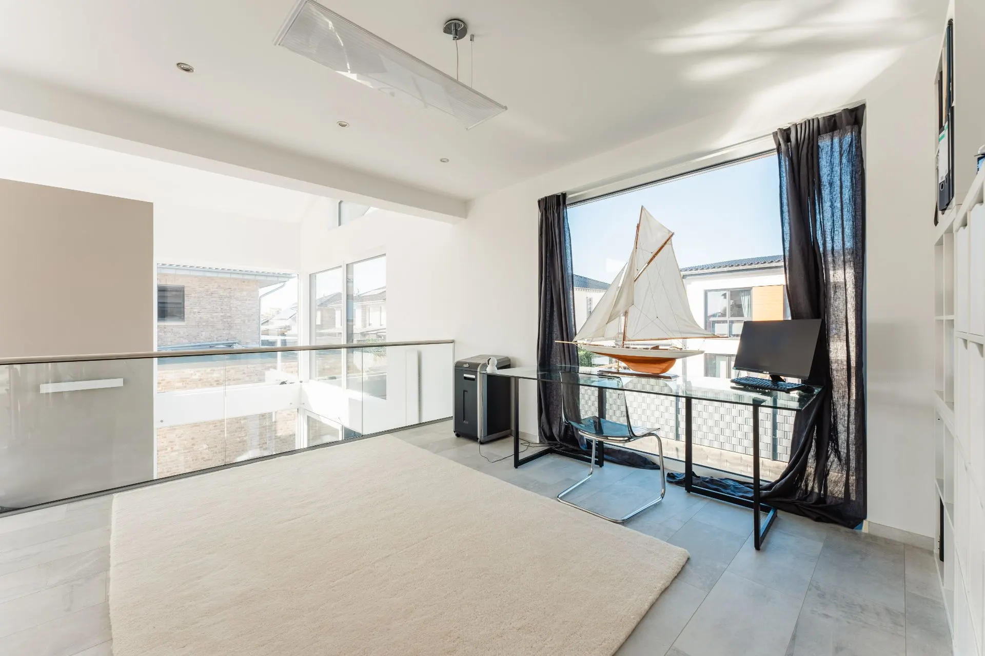 Helles Arbeitszimmer mit Glastisch, Stuhl, Modellsegelboot, großem Fenster und hellem Teppich.