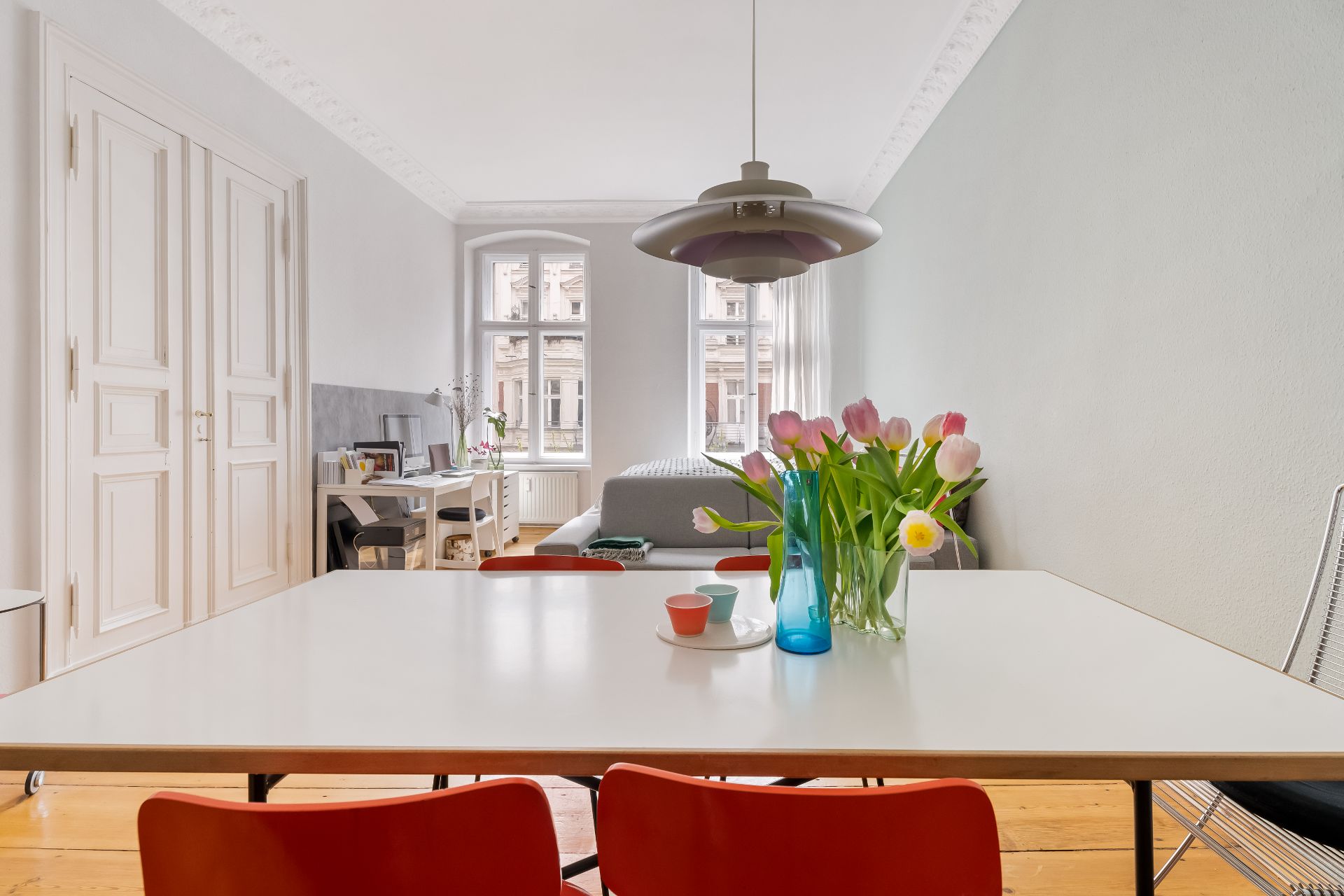 Heller Wohnraum mit Esstisch, roten Stühlen, Blumen und Blick auf Sofa und Schreibtisch.