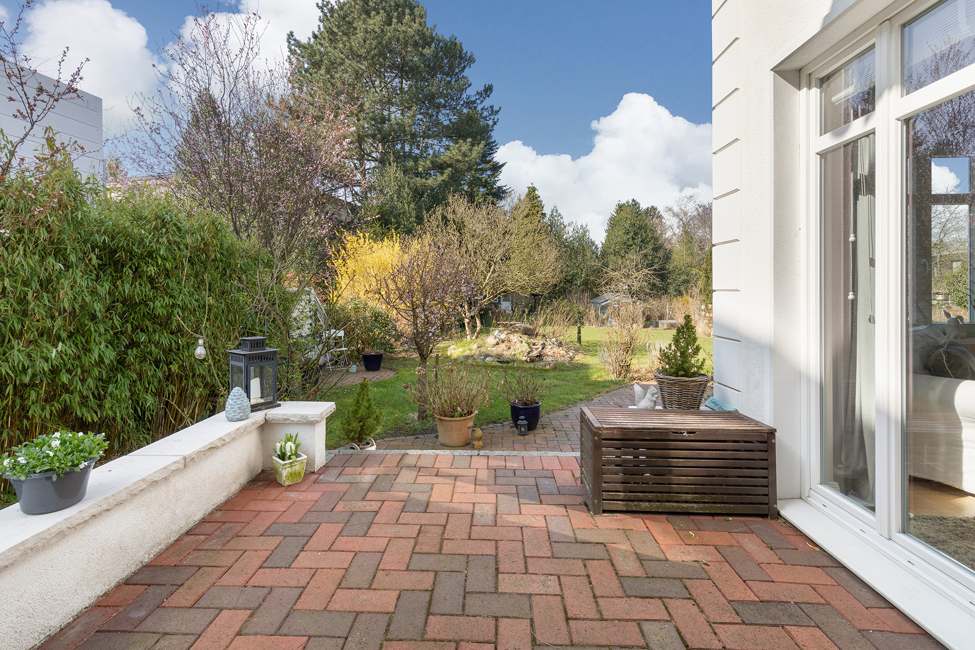 Gepflasterte Terrasse mit Pflanzen und Blick in einen grünen Garten mit Bäumen.