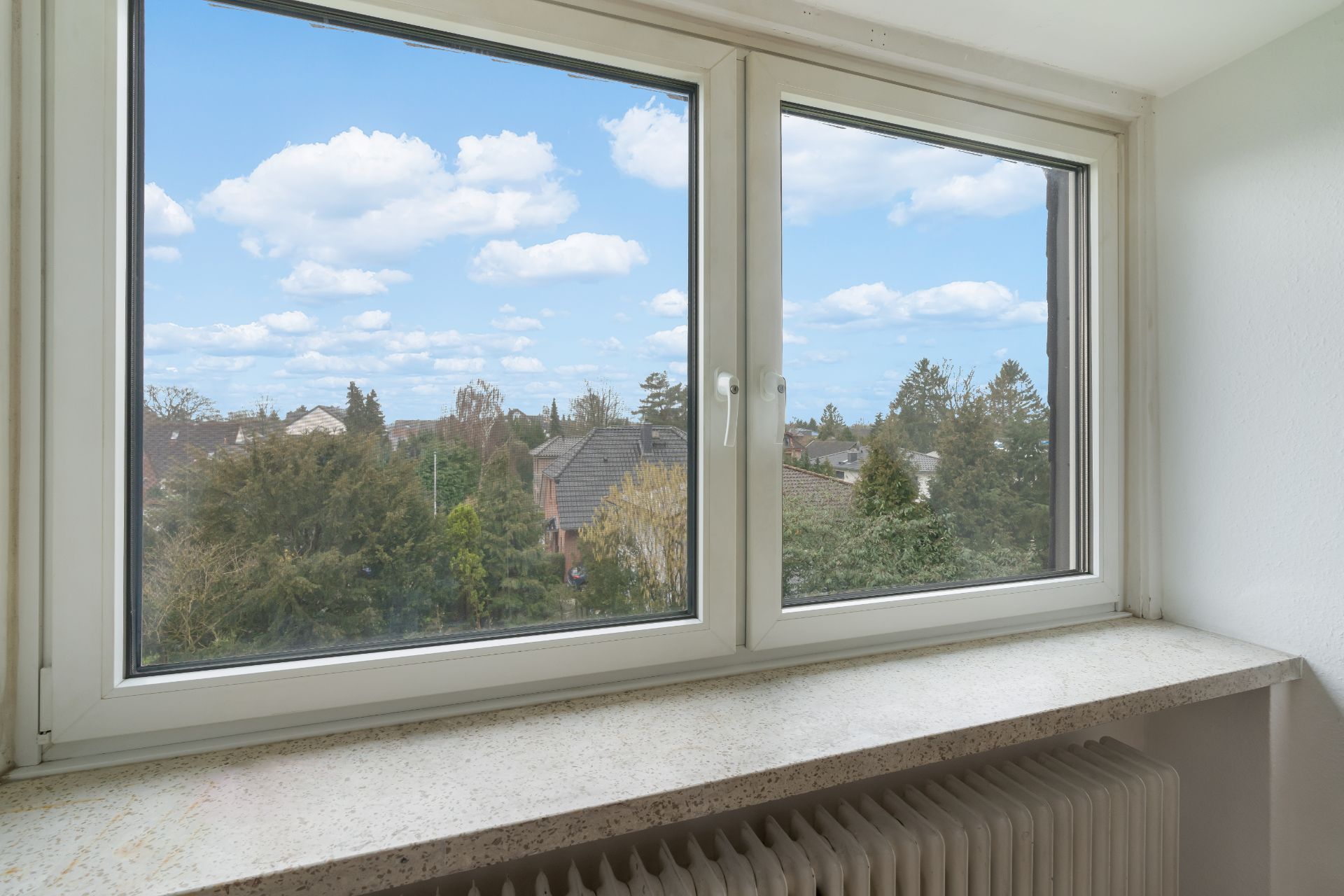 Blick aus einem Fenster auf Bäume und Wohnhäuser, mit Heizkörper unter der Fensterbank.