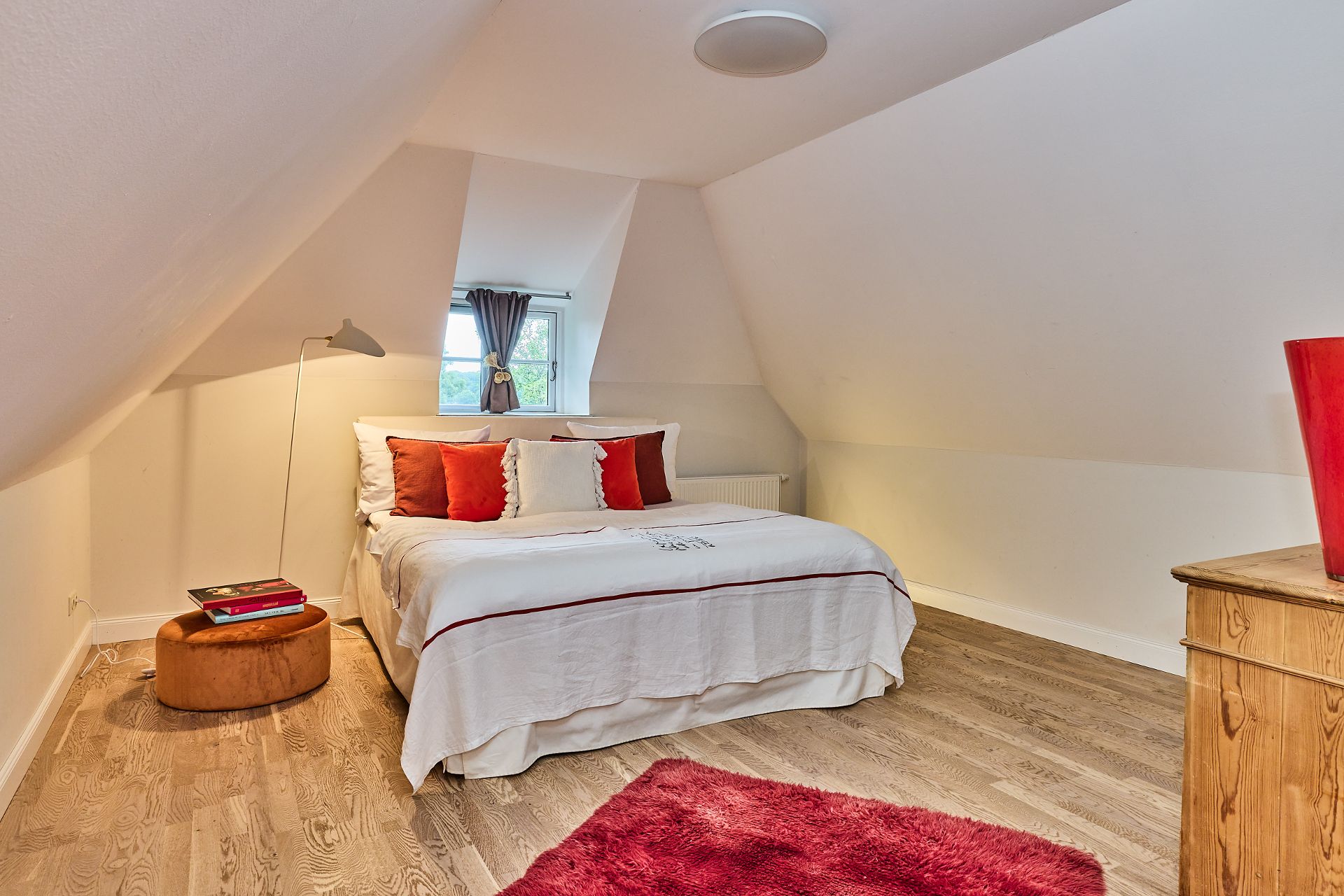Schlafzimmer mit Doppelbett, rotem Teppich, Nachttisch und Fenster mit Vorhang.