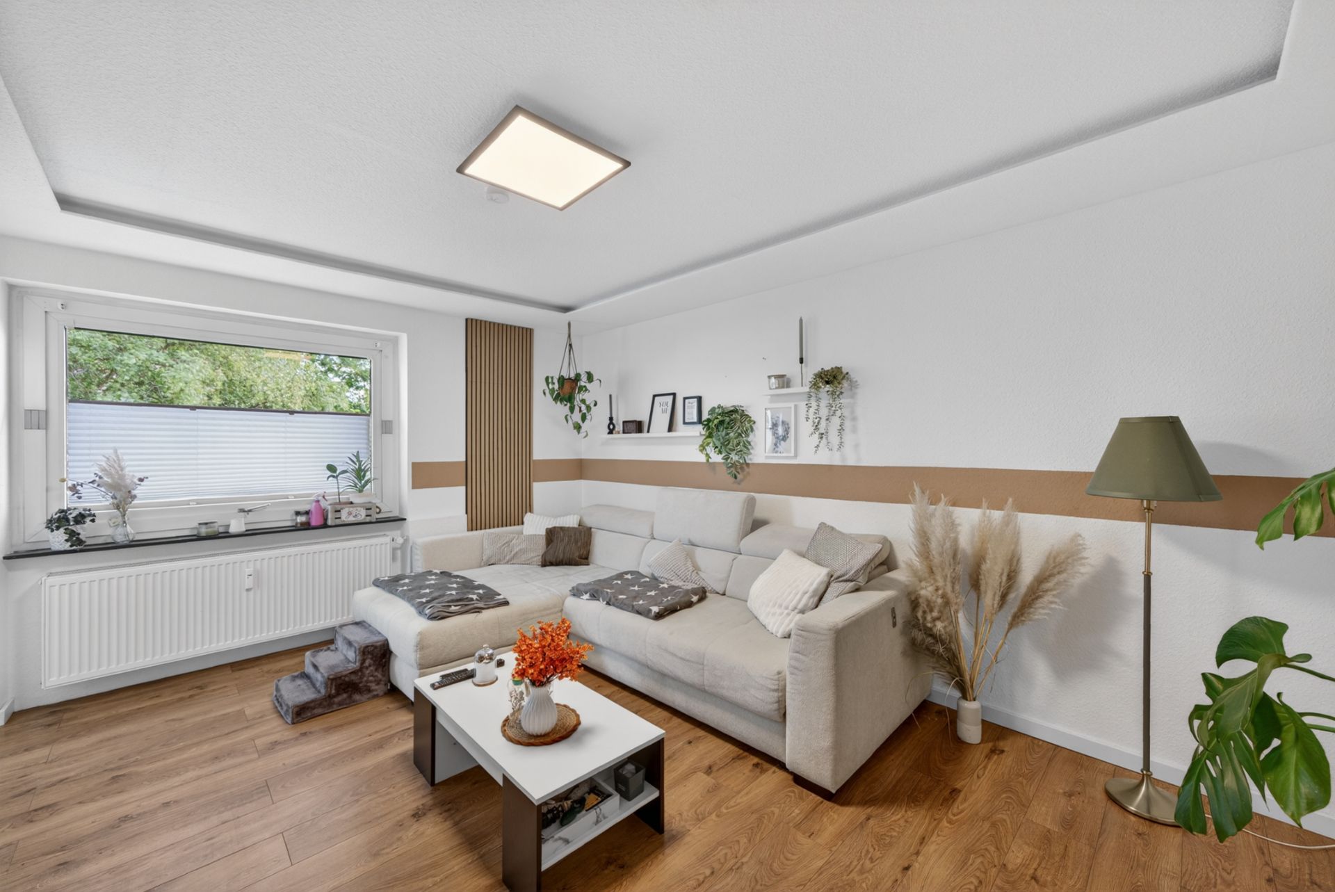 Helles Wohnzimmer mit Ecksofa, Couchtisch, großem Fenster und Pflanzen auf Holzboden.