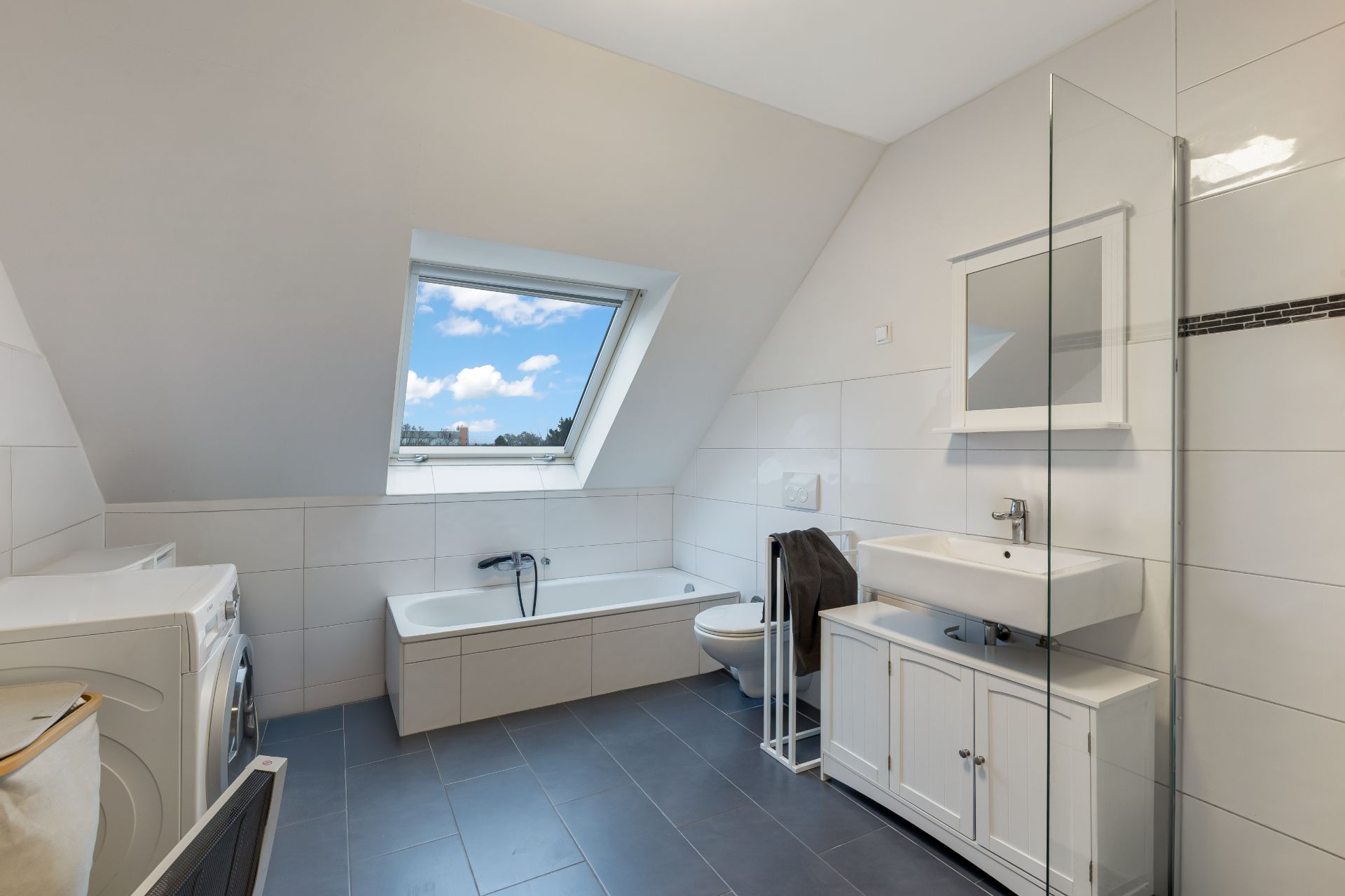 Helles Badezimmer mit Badewanne, Waschbecken, Toilette, Waschmaschine und großem Dachfenster.