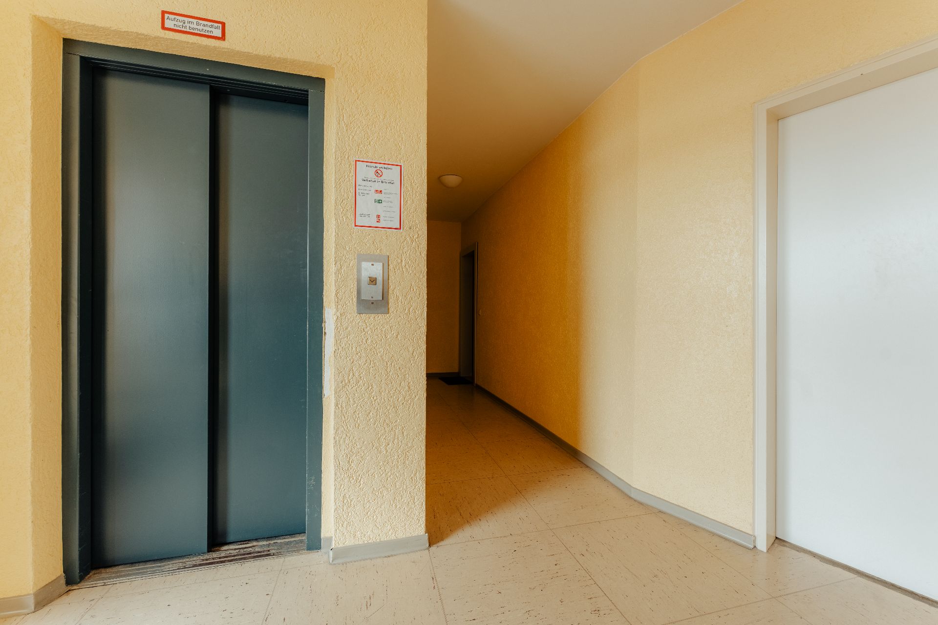 Ein Aufzug und ein Flur mit gelben Wänden in einem Wohnhaus sind sichtbar.