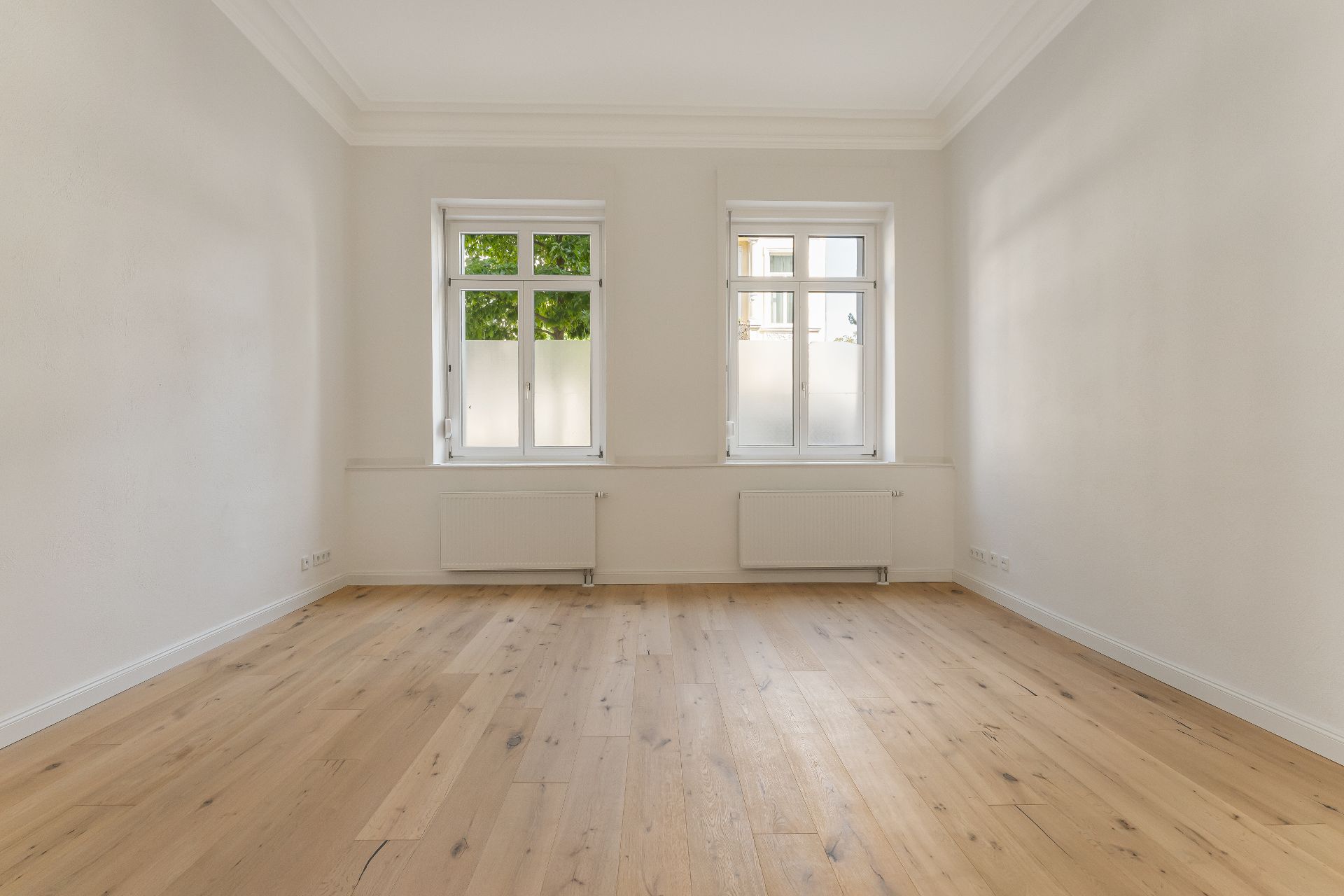 Leerer Raum mit hellem Holzfußboden, weißen Wänden und zwei Fenstern.