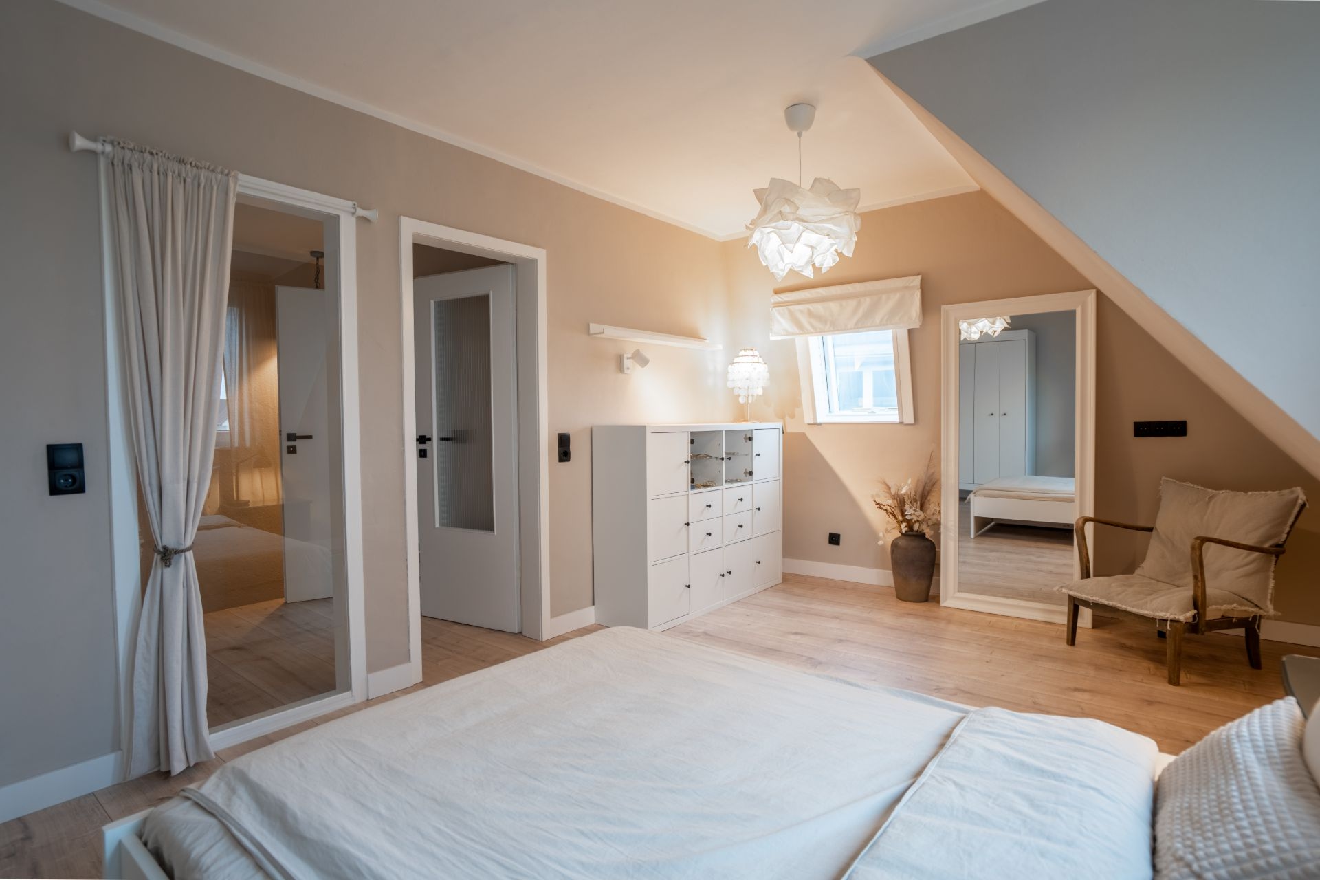 Helles Schlafzimmer mit Bett, Kommode, Sessel und großem Spiegel an der Wand.
