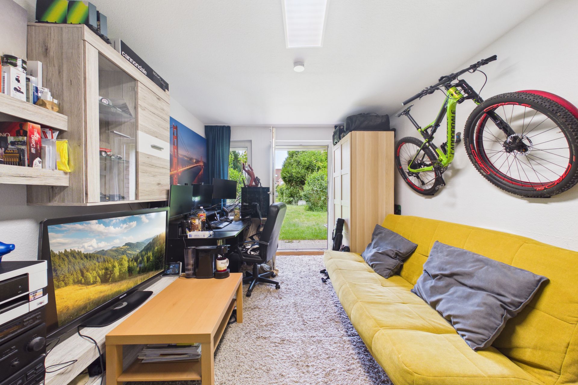 Wohnzimmer mit gelbem Sofa, Fernseher, Schreibtisch und Fahrrad an der Wand, Tür führt zum Garten.