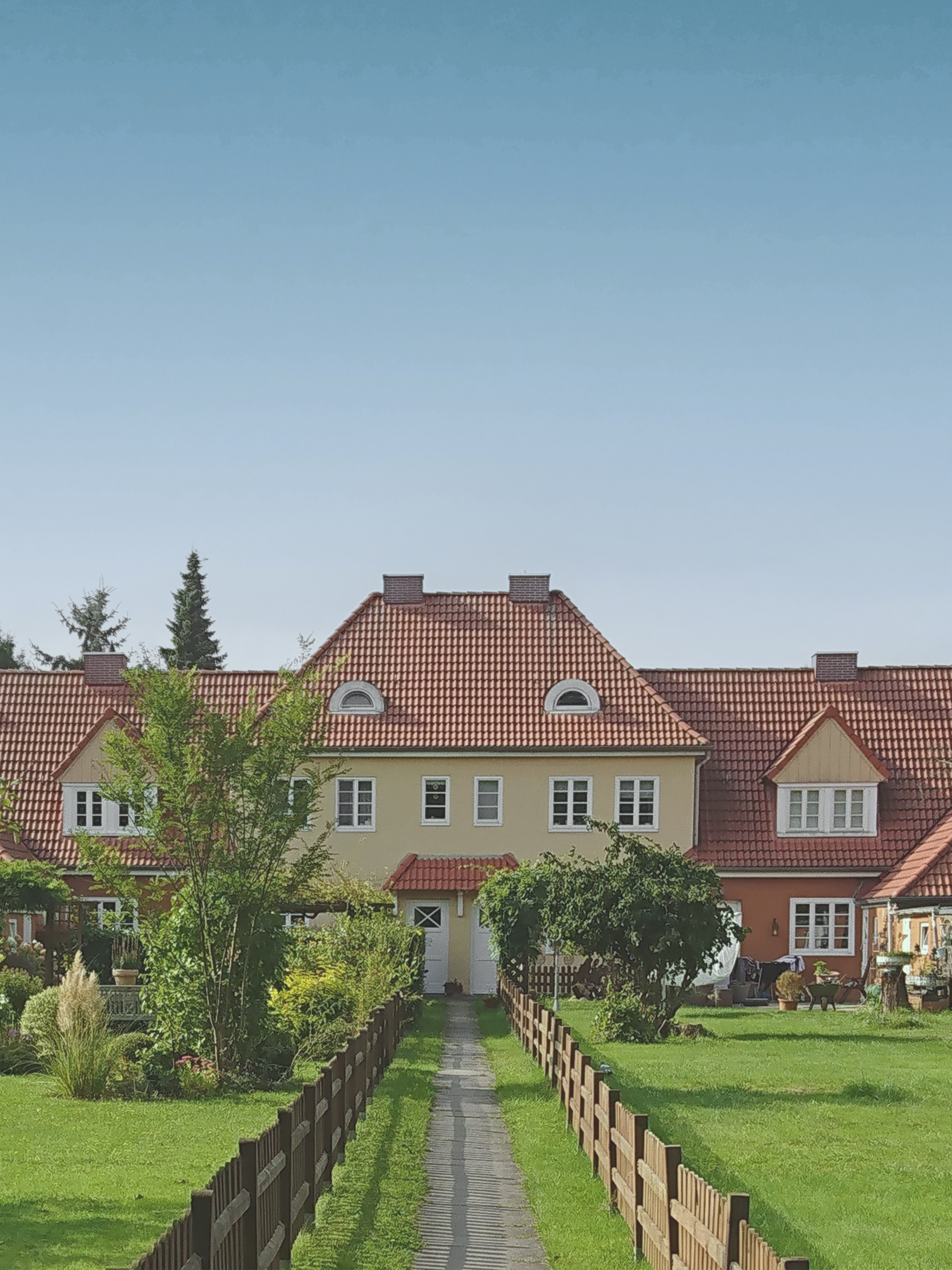 Reihenhaus mit rotem Dach, Garten und Holzzaun im Vordergrund bei klarem Himmel.