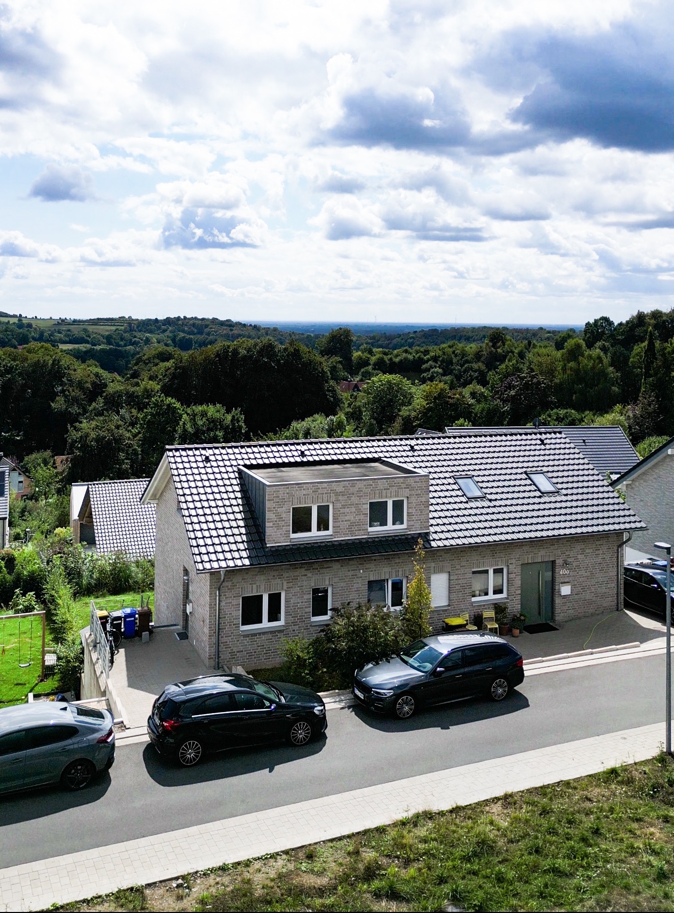 Modernes Einfamilienhaus mit grauem Dach, Autos parken davor, grüne Landschaft im Hintergrund.