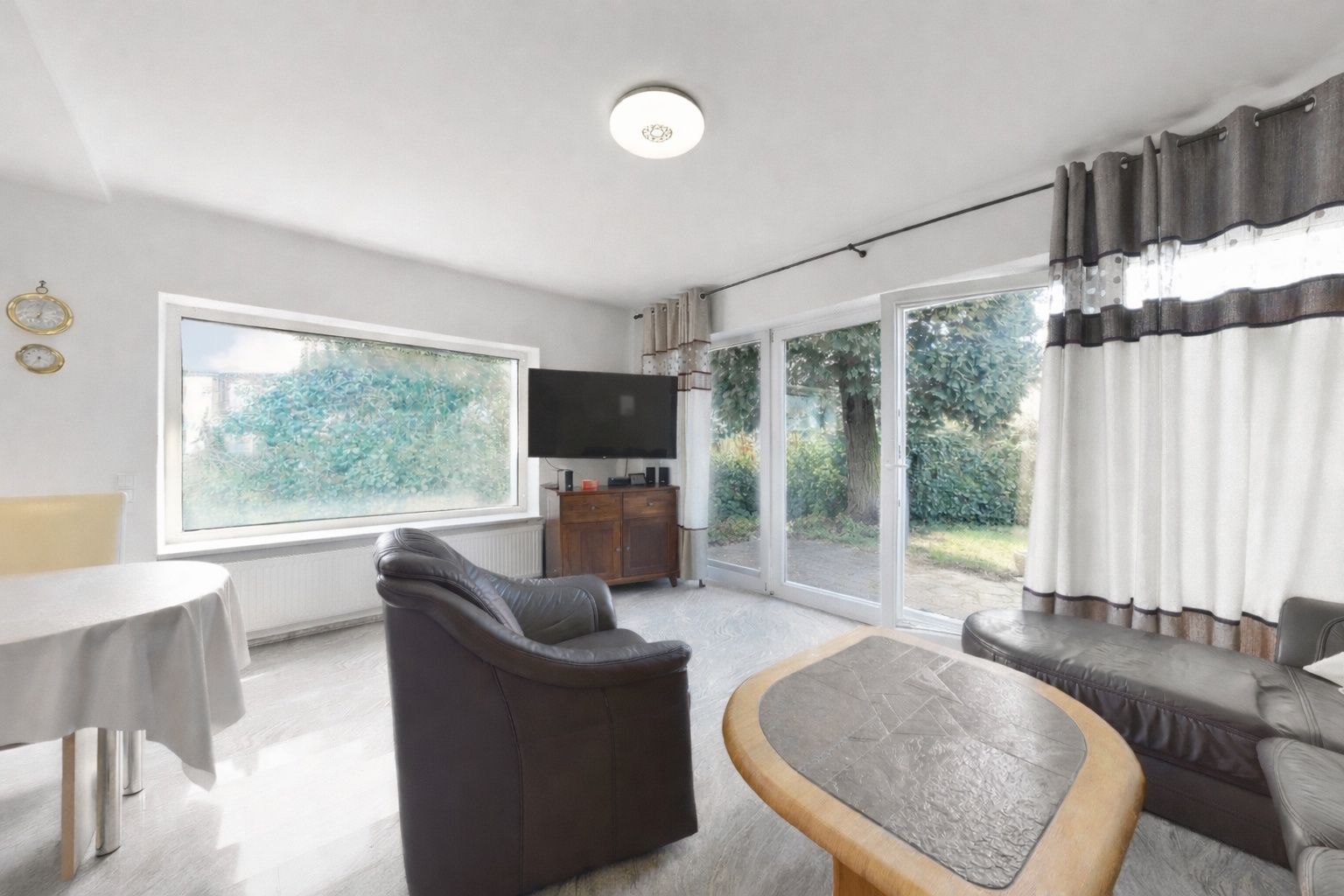 Helles Wohnzimmer mit Sofa, Sessel, Couchtisch, Fernseher und großen Fenstern zum Garten.