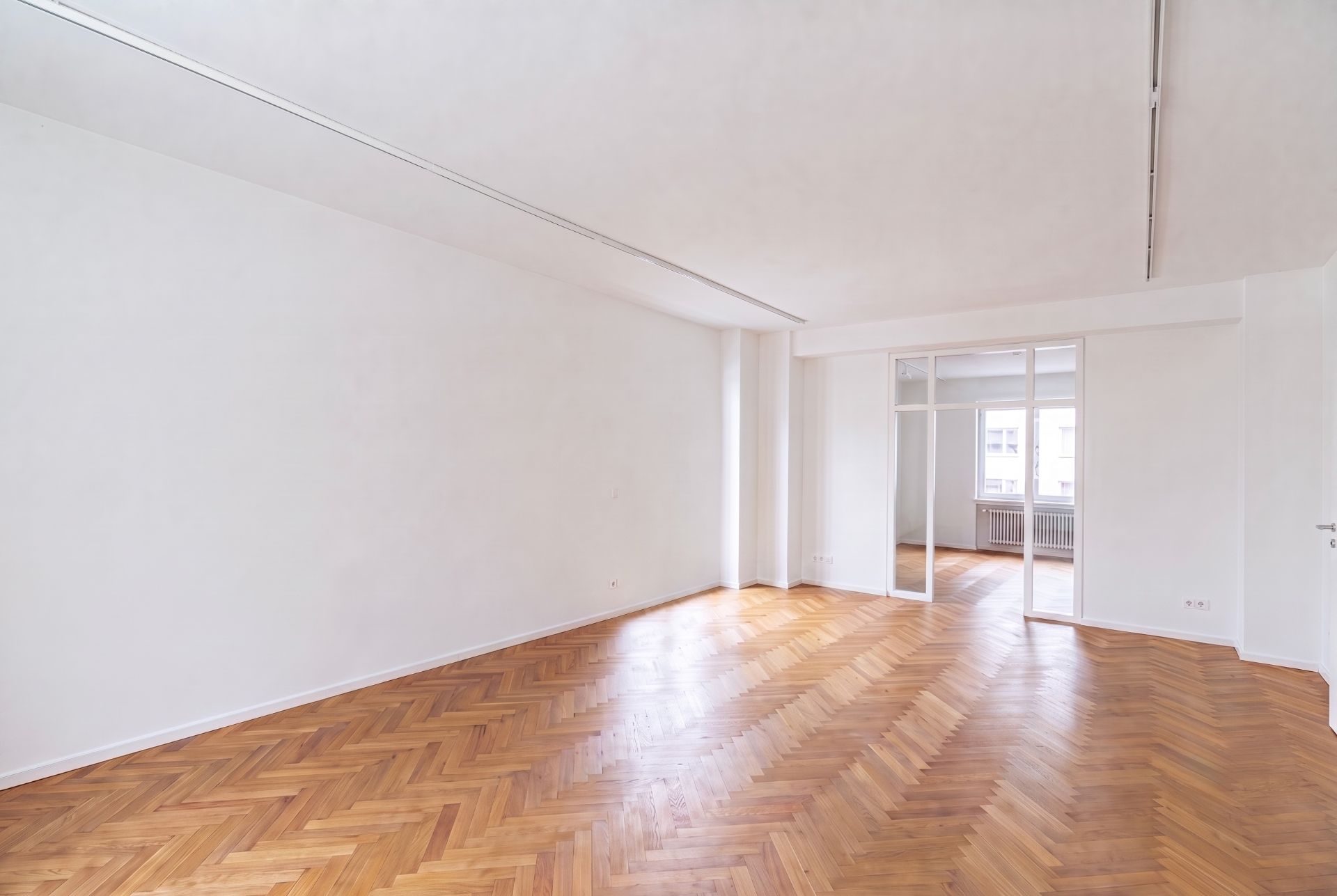 Leerer Raum mit Parkettboden und weißen Wänden, große Fenster und Glastür zur nächsten Etage.